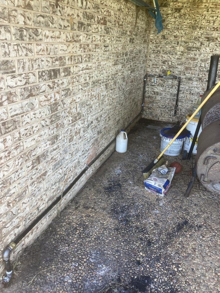 Brick walled outdoor area with pipes on the floor, cleaning supplies, and a partially visible container.