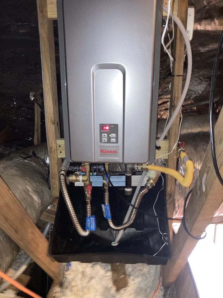 Rinnai tankless water heater, silver, installed in an attic. Plumbing and gas lines visible, wood framing.