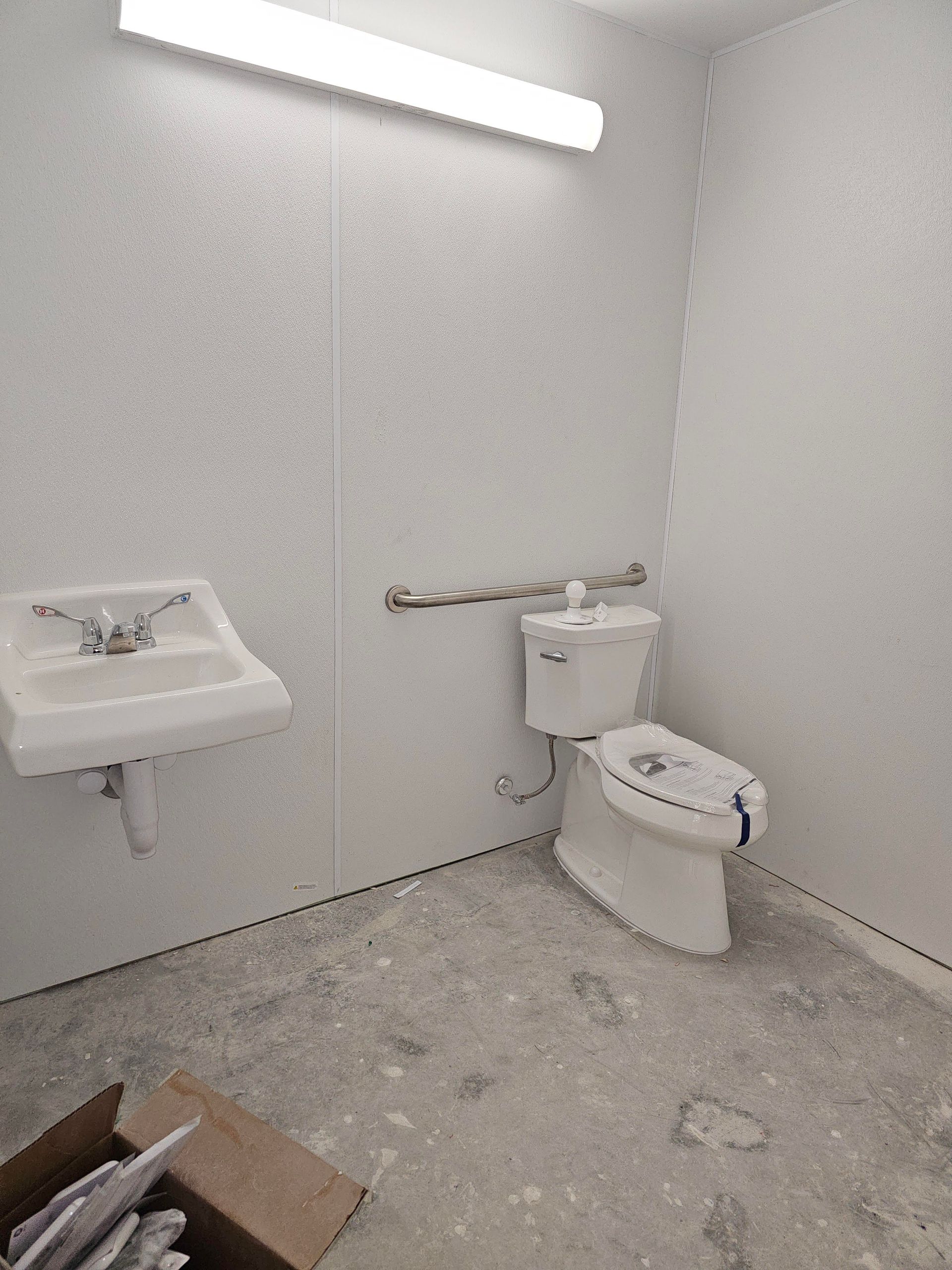 Accessible restroom with sink, toilet, and grab bar. White walls, gray floor, overhead light.