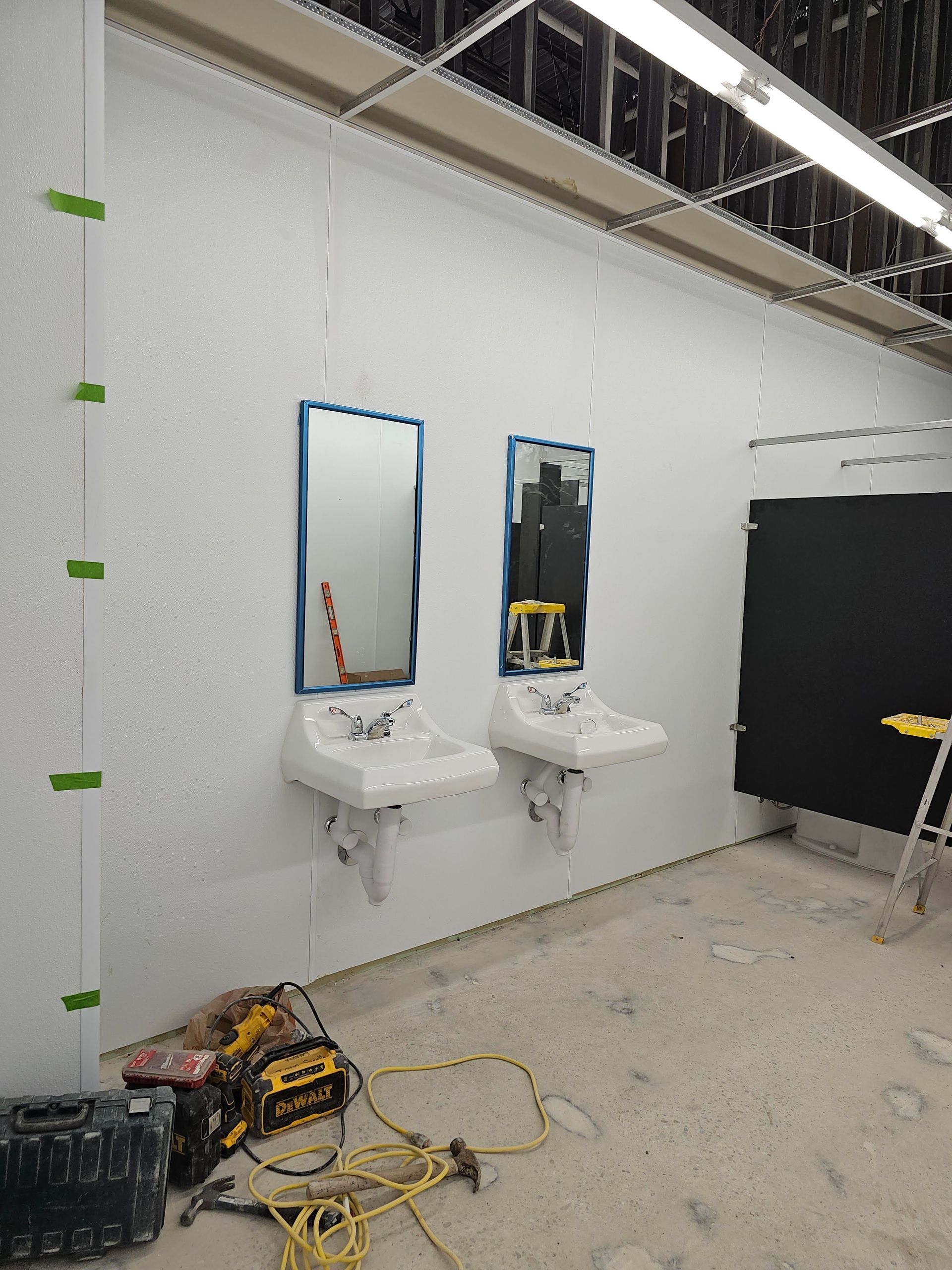 Unfinished restroom with two sinks, mirrors, and black stall. Construction tools are on the floor.