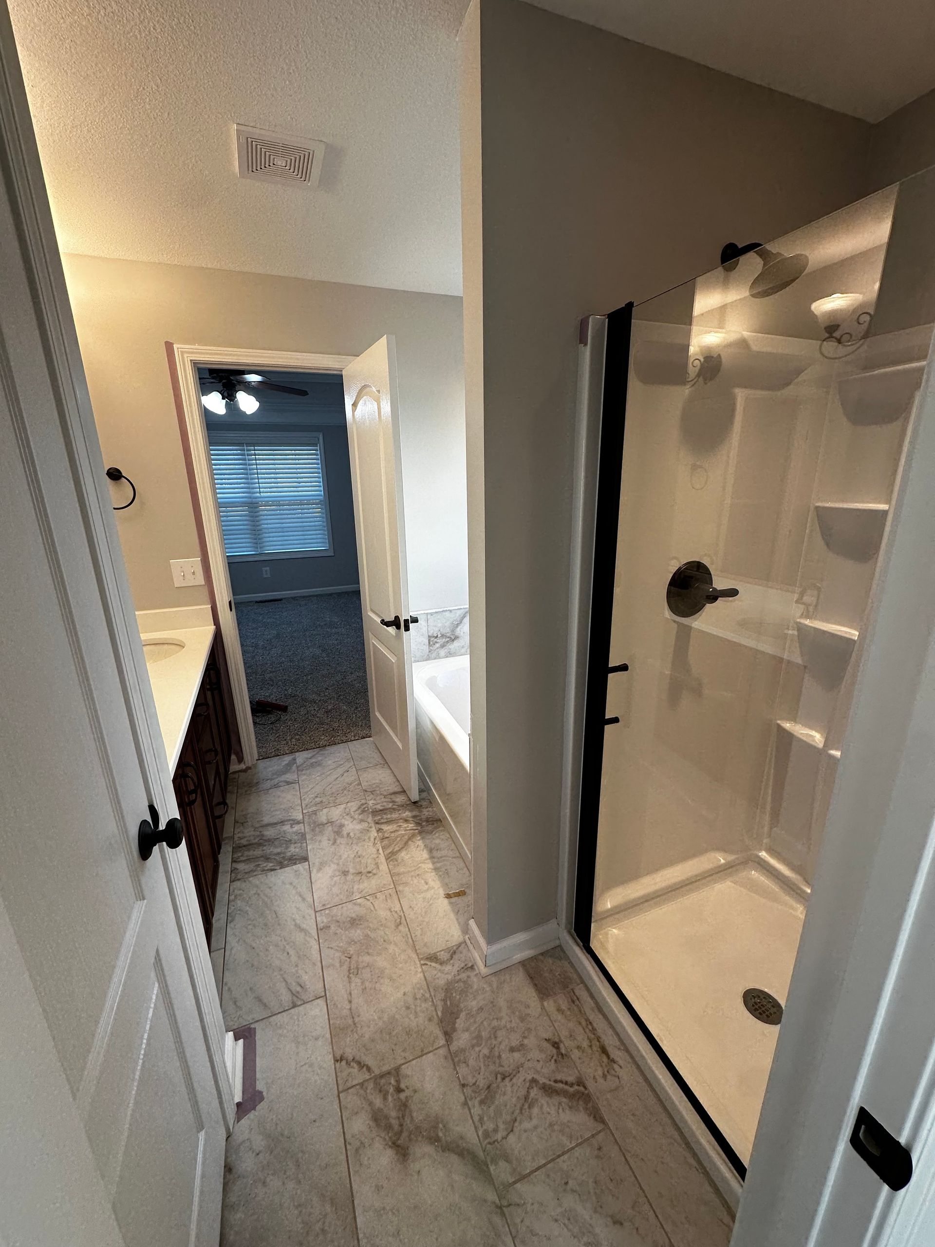 Bathroom with shower, vanity, and doorway to a bedroom, featuring light gray walls, white trim, and a tile floor.