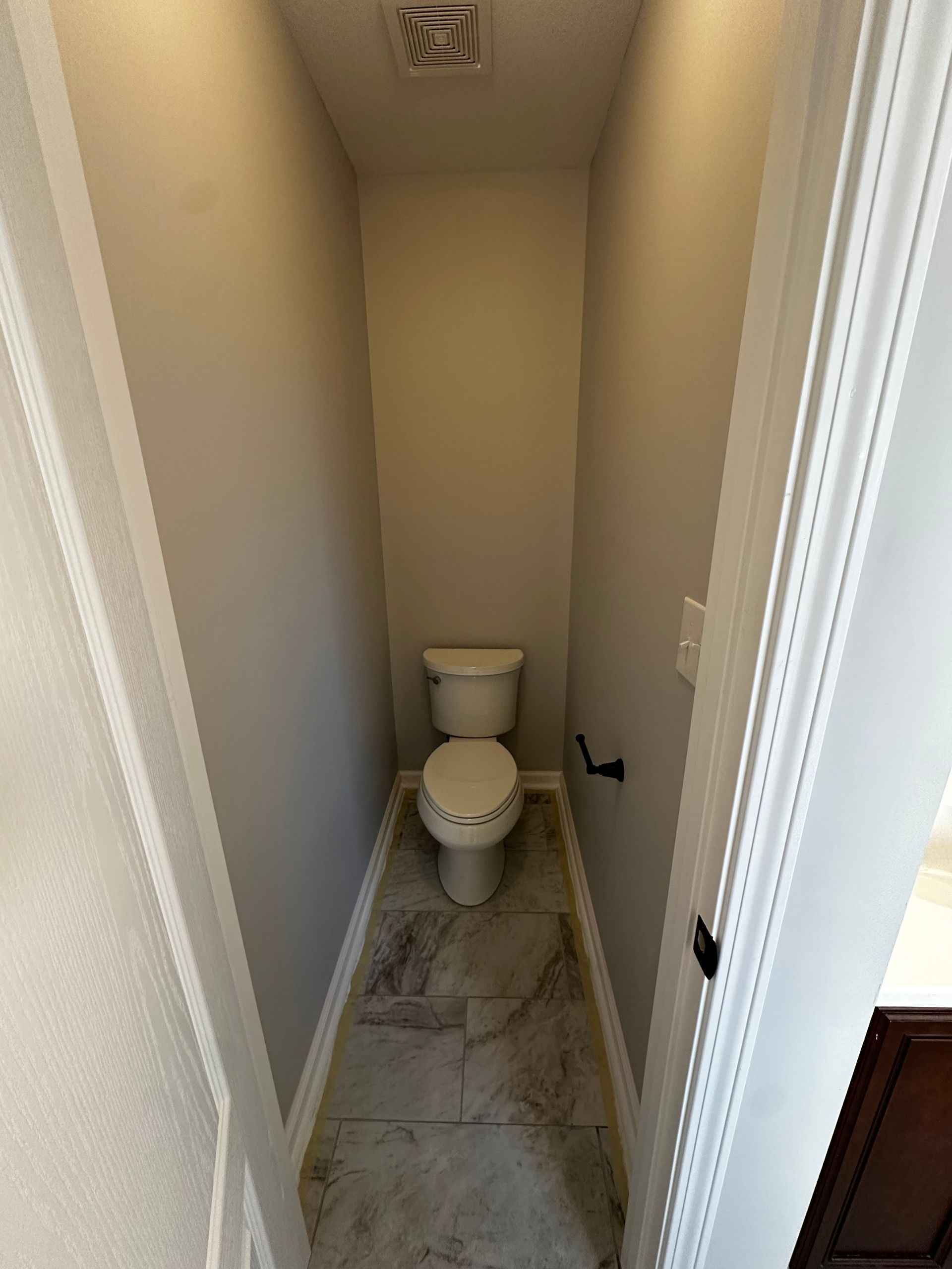 Narrow bathroom with a toilet, light gray walls, and light-colored tiled floor.
