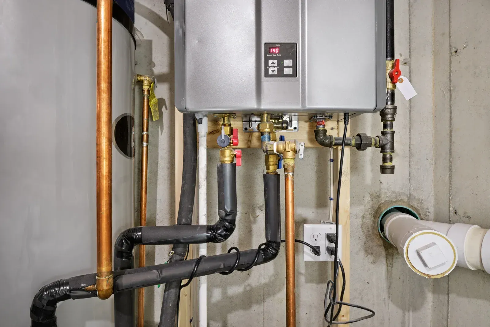 A wall-mounted, silver tankless water heater with copper pipes in a basement setting.