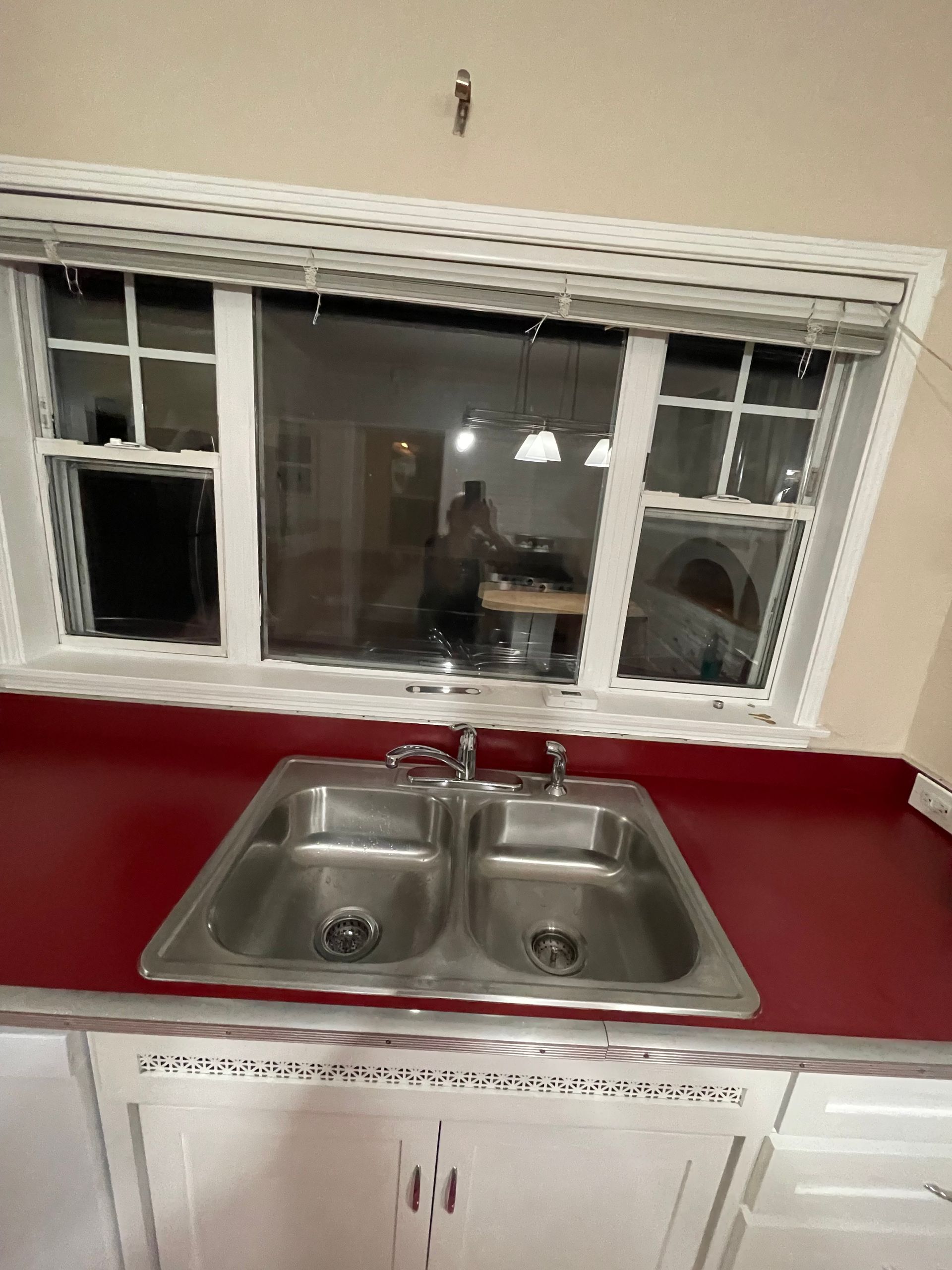 Kitchen sink in front of a window, red countertop, white cabinets.