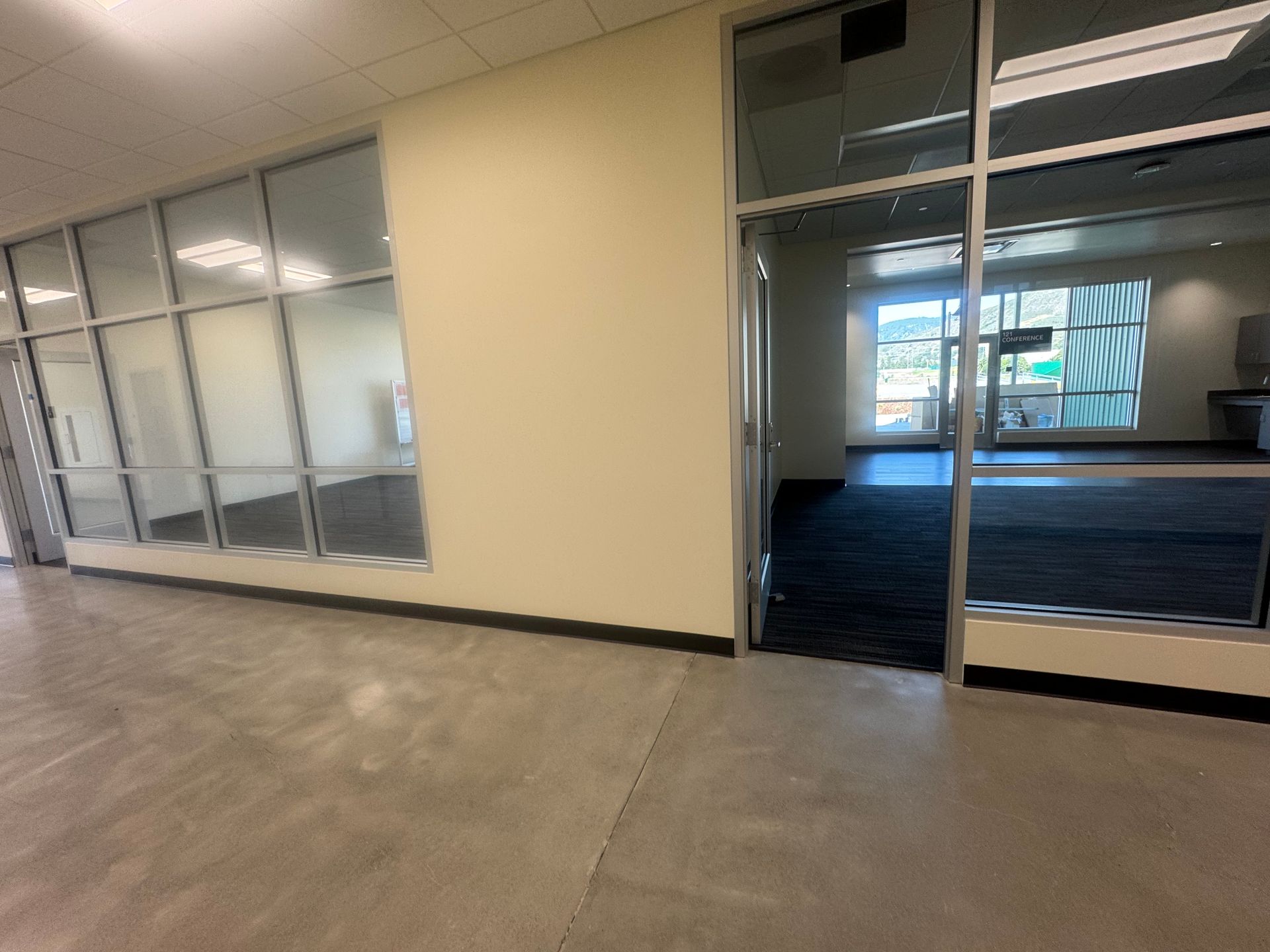 Interior view: corridor with glass walls, cream wall, and an open doorway to a dark room.
