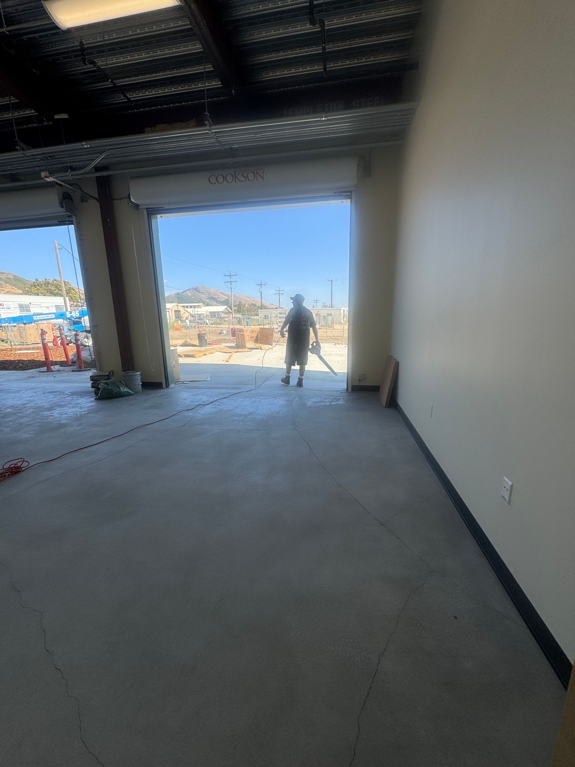 Interior of a building with an open garage door, worker standing outside, concrete floor, neutral walls.