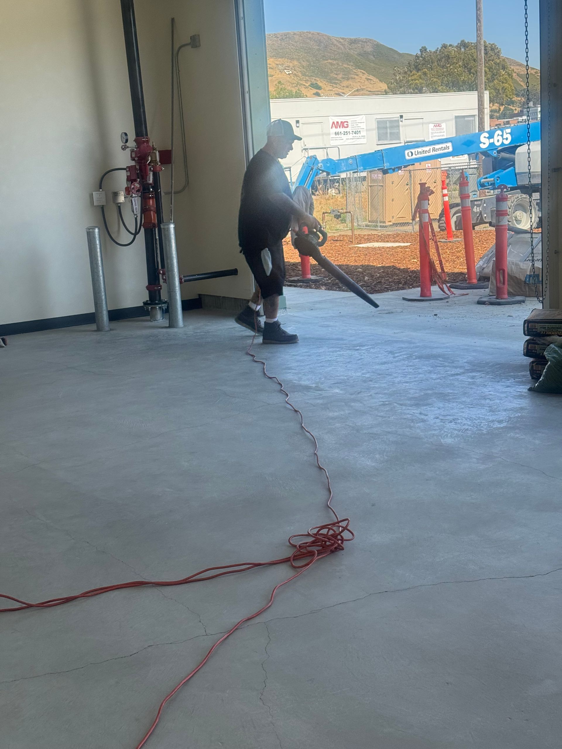 Person in dark clothing uses a leaf blower on a concrete floor. Red cord in foreground, building and mountain in background.