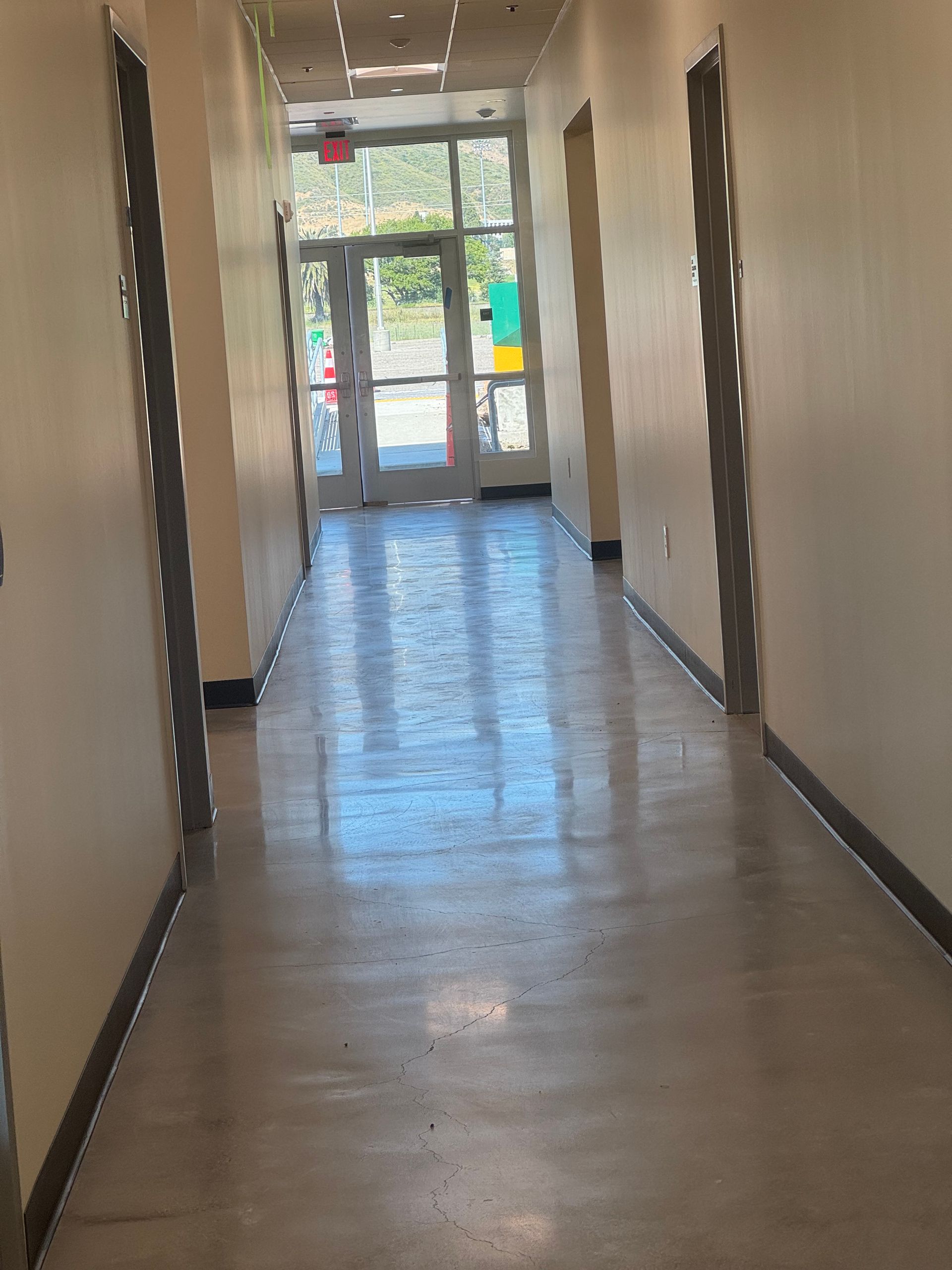 Long hallway with gray floor, off-white walls, doors on each side, and an exterior door at the end.
