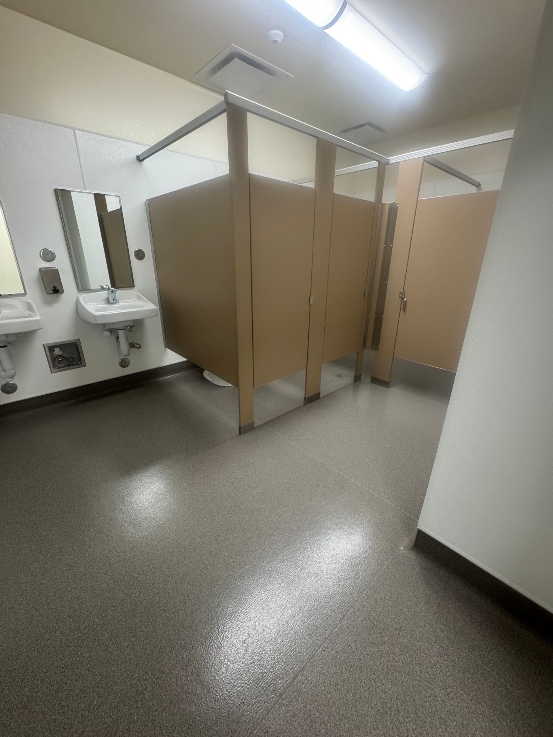 Empty public restroom with tan stall doors and speckled gray floor.