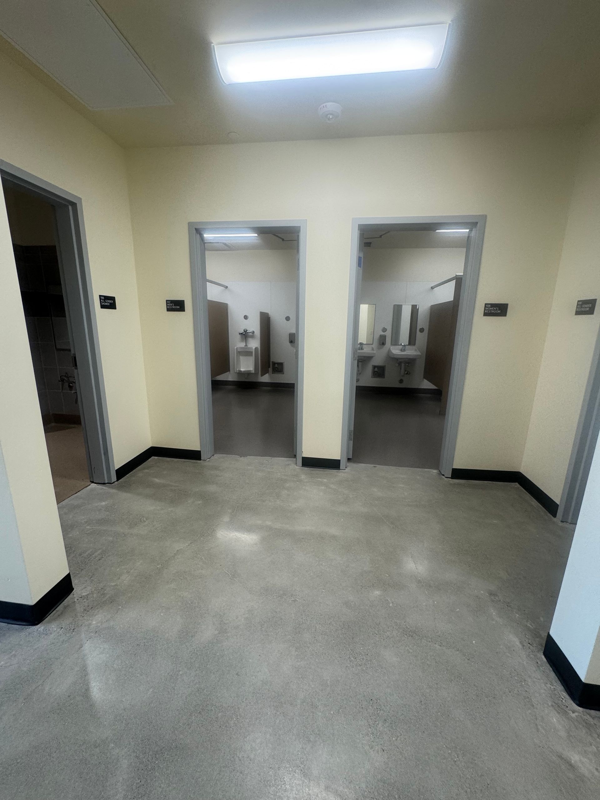 Hallway with three open doorways. Two lead to restrooms, with sinks and stalls visible. Light walls, gray floor.