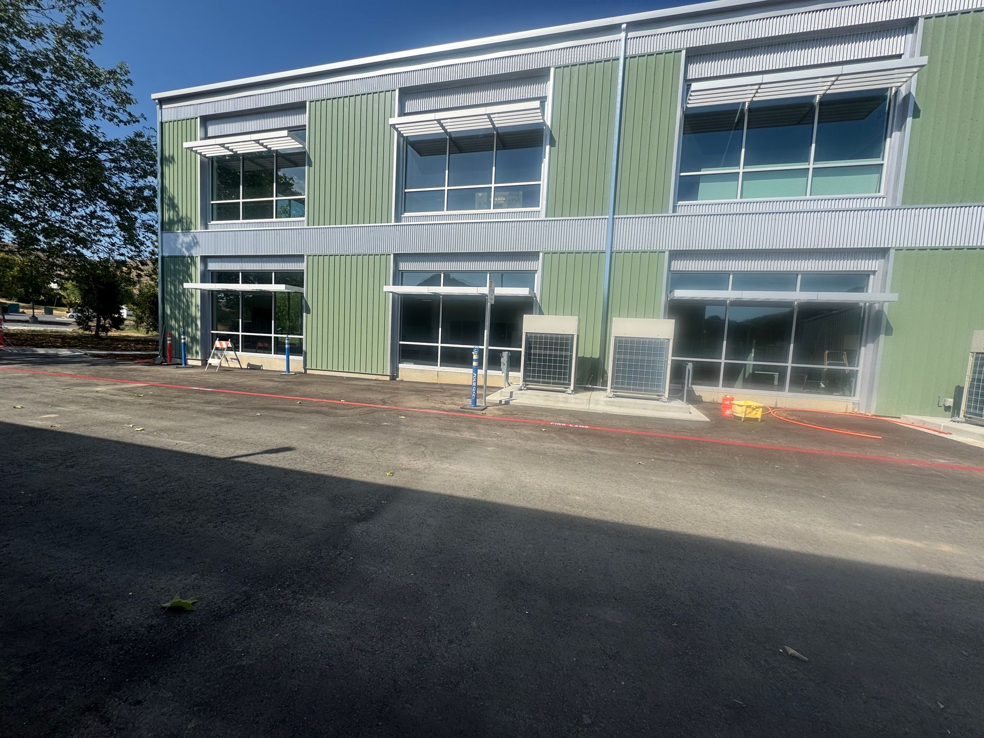Green and gray two-story building with large windows, construction zone in front; asphalt in foreground, blue sky.