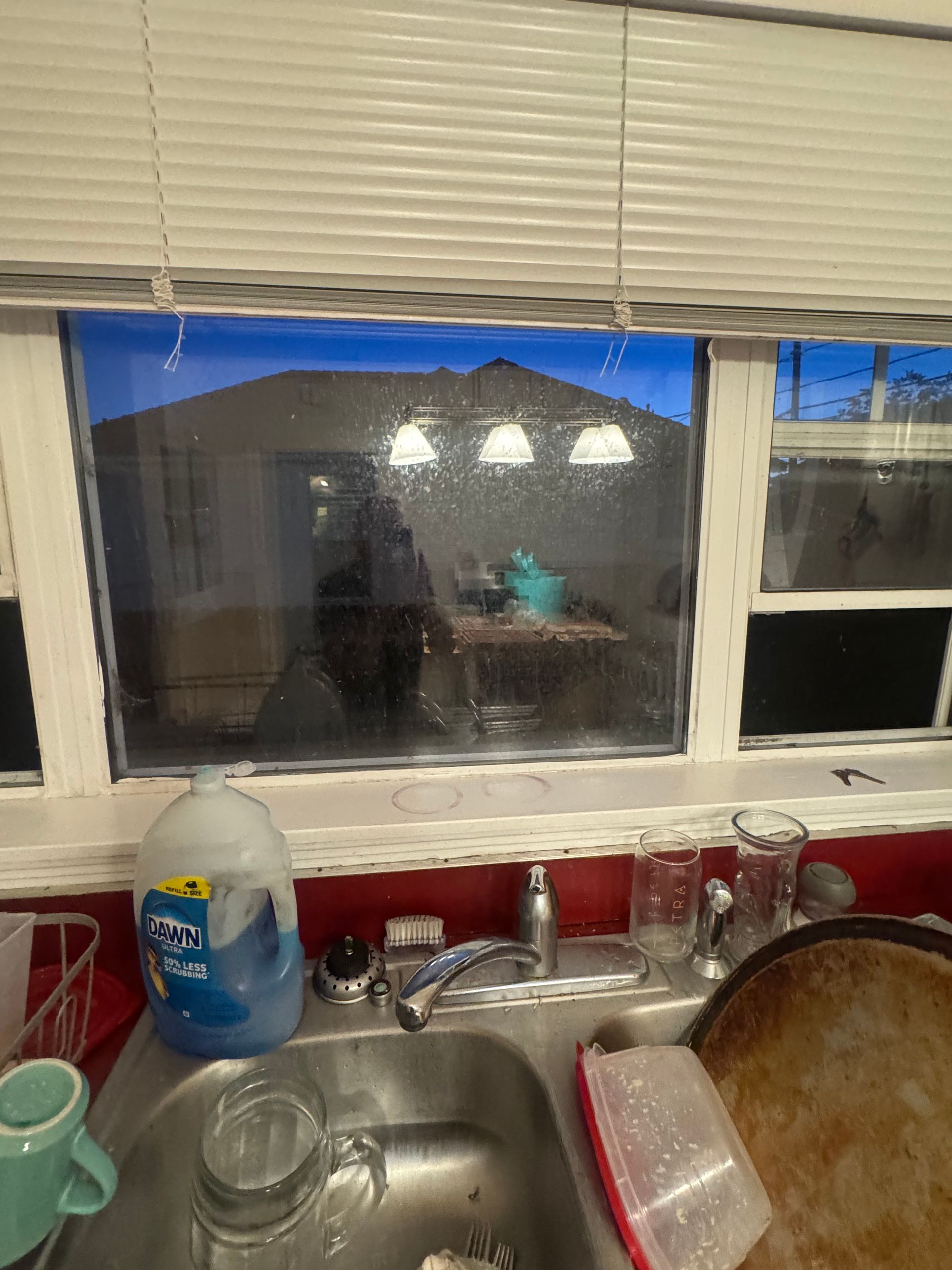Kitchen sink by a window, overlooking a backyard. Dirty dishes, soap, and outdoor lights are visible.