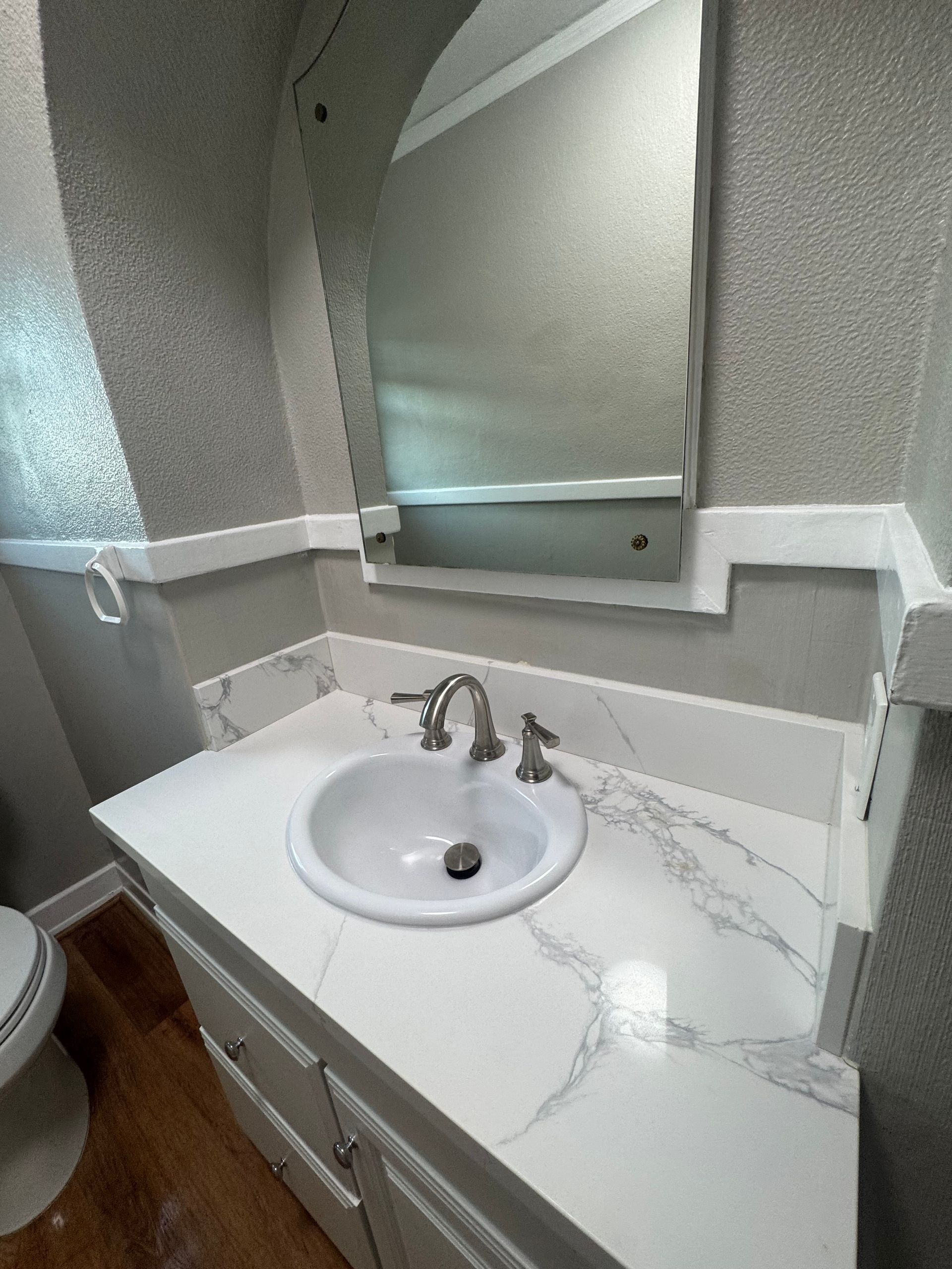 Bathroom with white countertop, sink, and mirror; gray speckled wallpaper and wood floor.