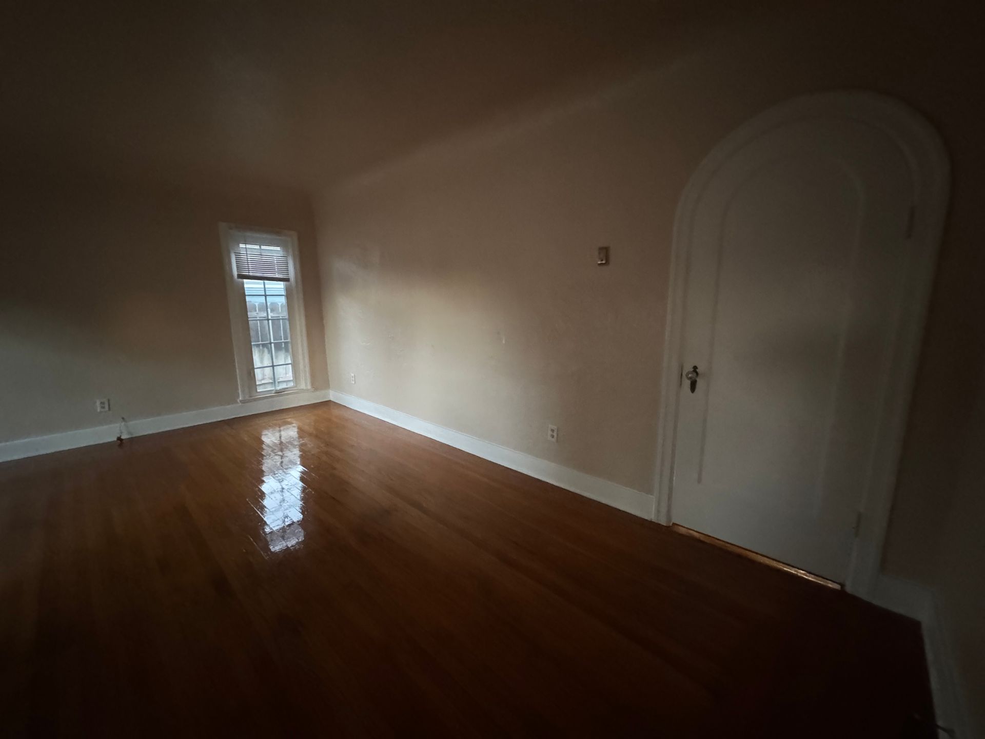 Empty room with hardwood floors, arched door, small window, and light-colored walls.