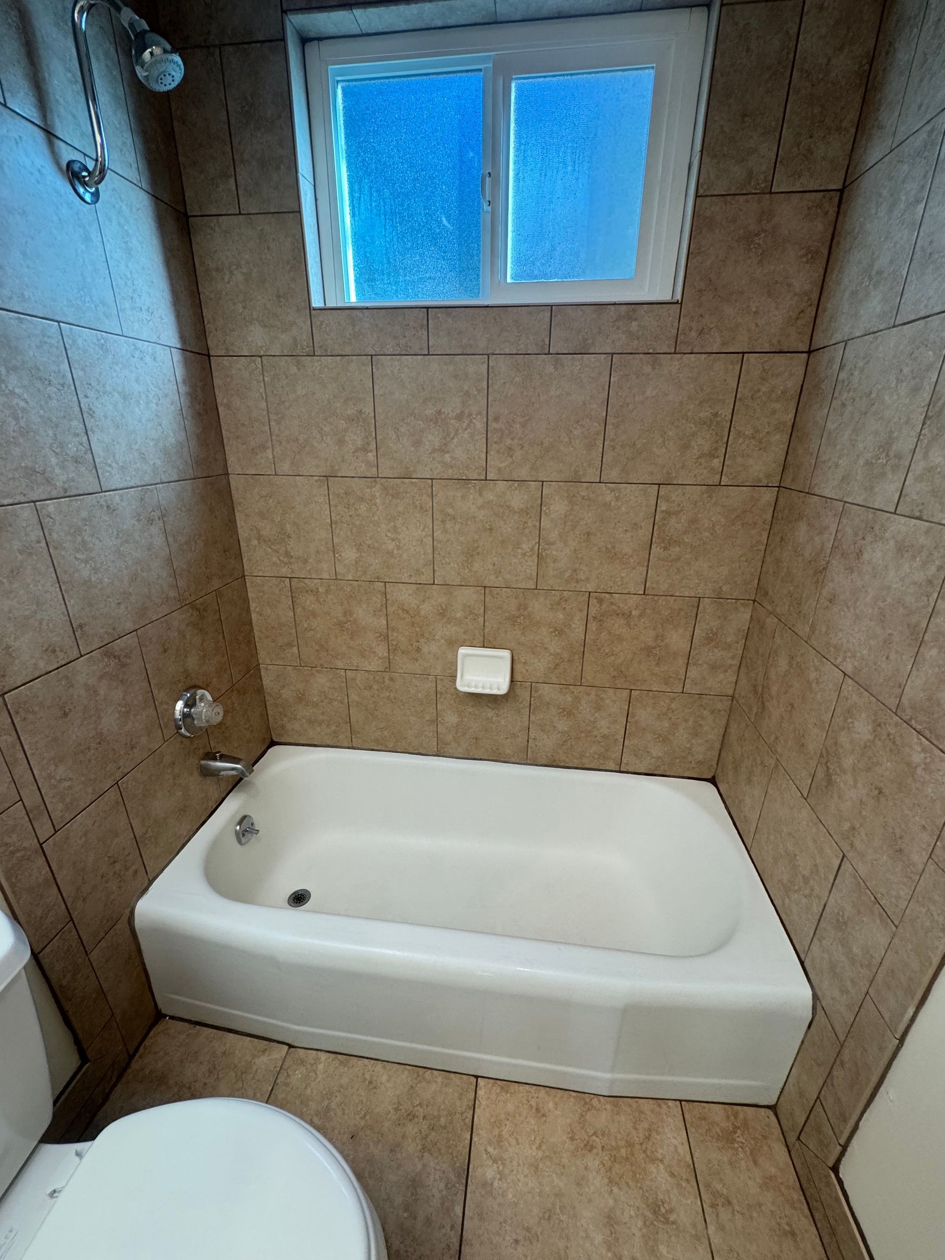 Small bathroom with a white bathtub, toilet, and light brown tiles. A window is above the tub.