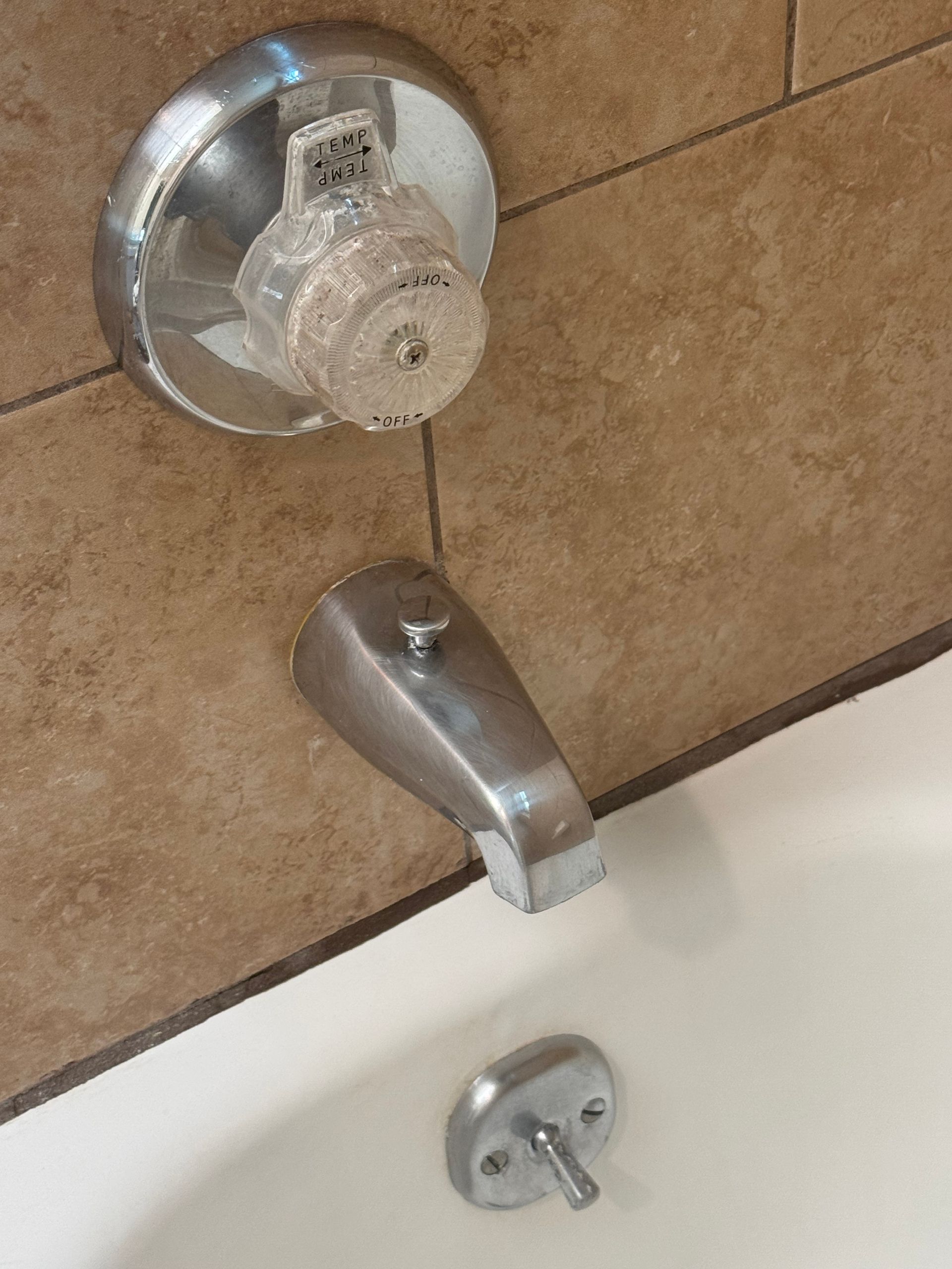 Tub faucet with clear handle and drain stopper in a beige tiled bathroom.