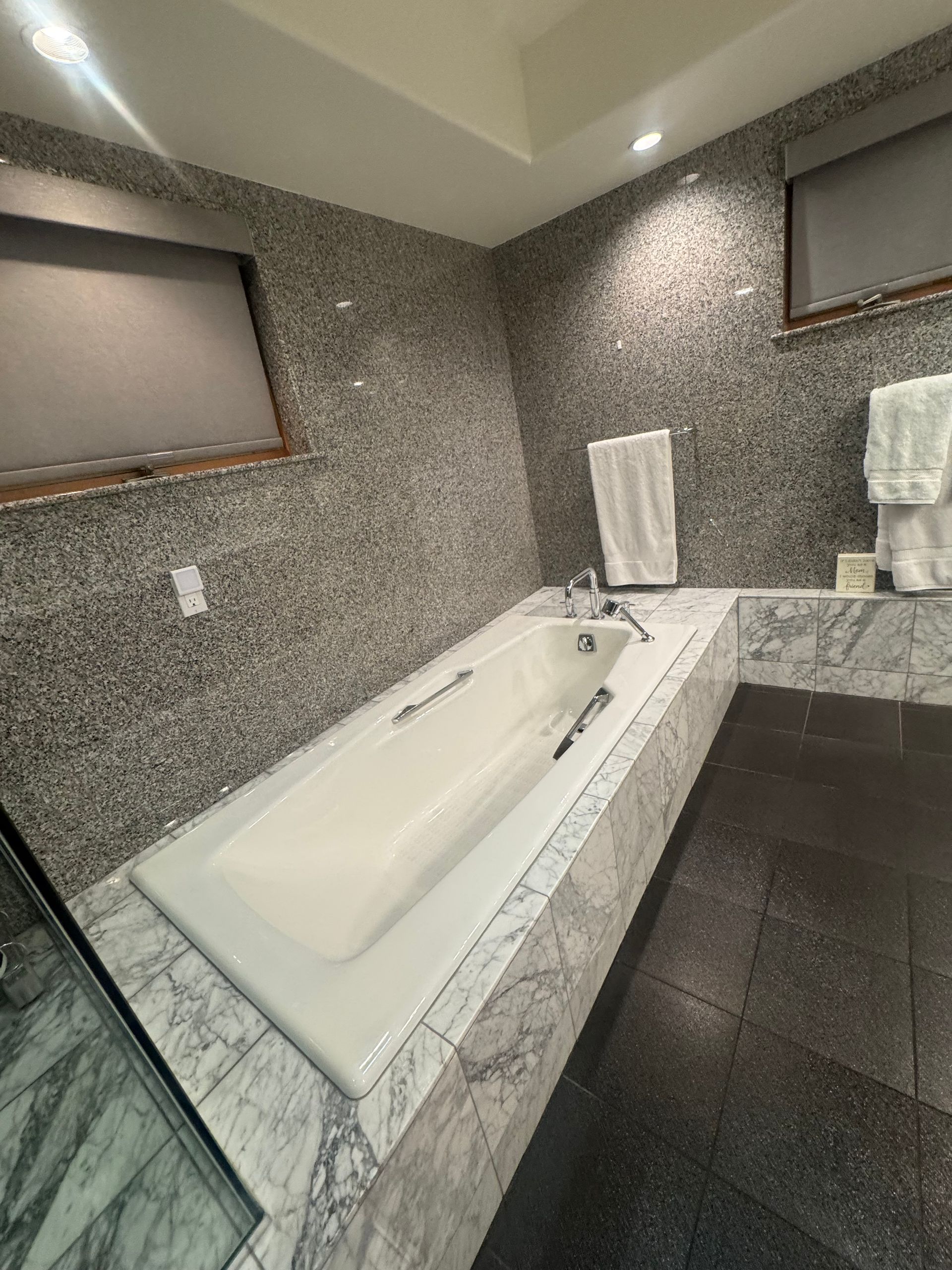 Bathroom with a rectangular bathtub, gray mosaic tile walls, and stone surround. Two towels are hanging.