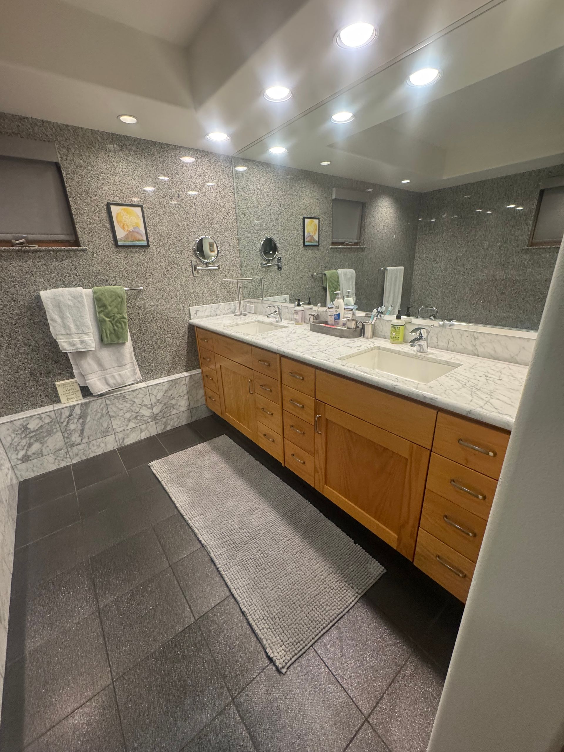 Bathroom with wood cabinets, marble countertop, and dark floor. A large mirror and mosaic tile wall are visible.