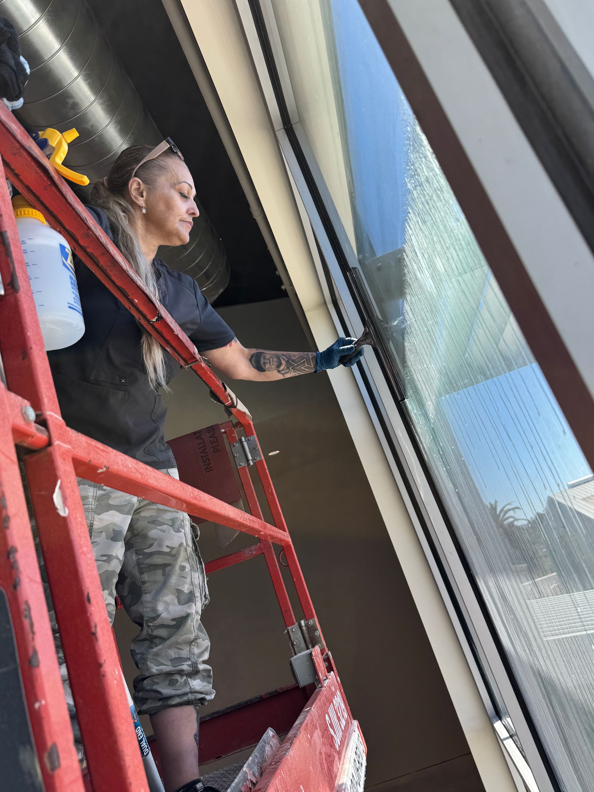 Woman on a lift cleaning a large window with spray bottle, squeegee. Outdoors, daytime.