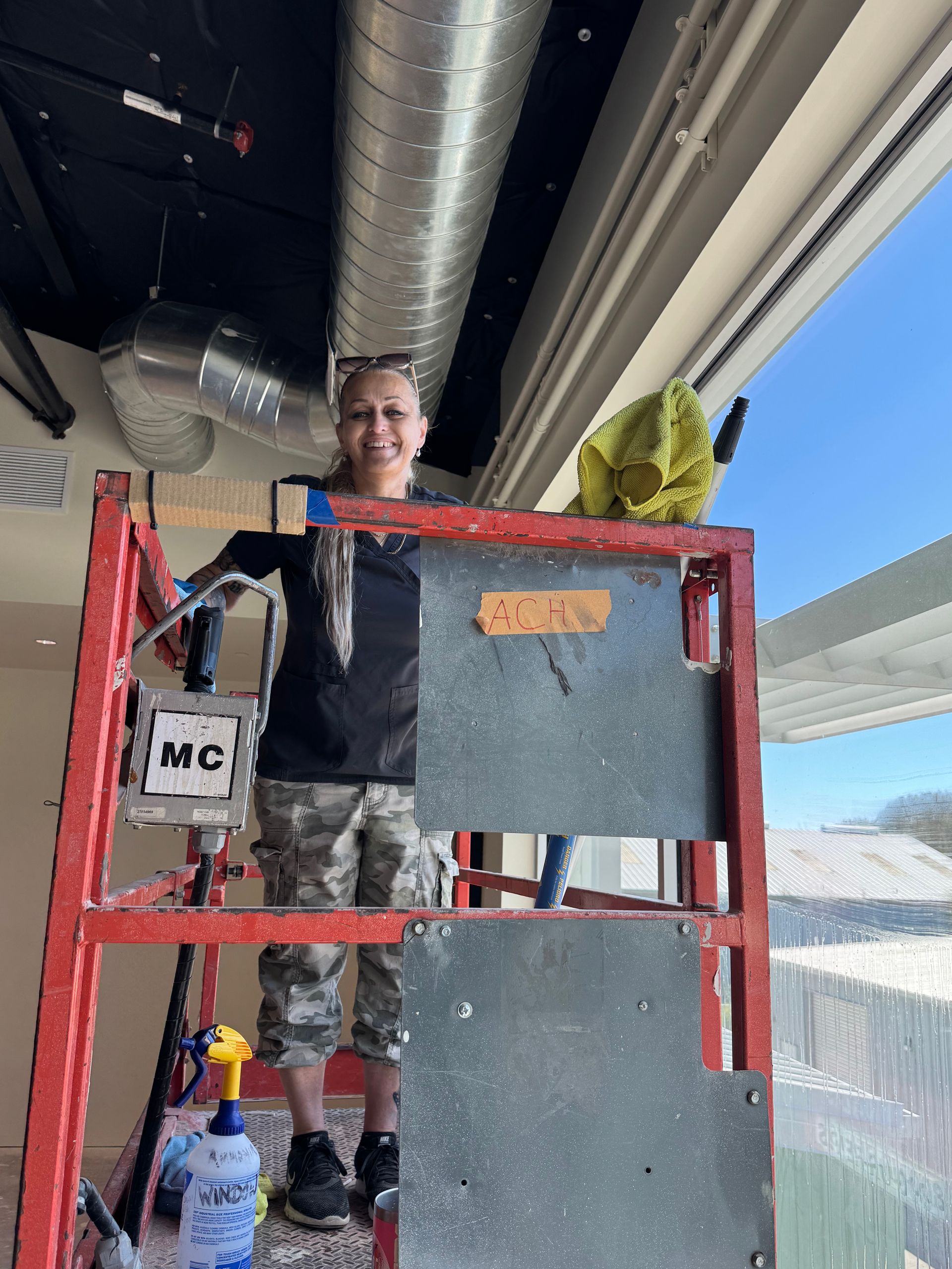 Woman in camouflage pants on a lift working near windows. Inside setting with silver ductwork.