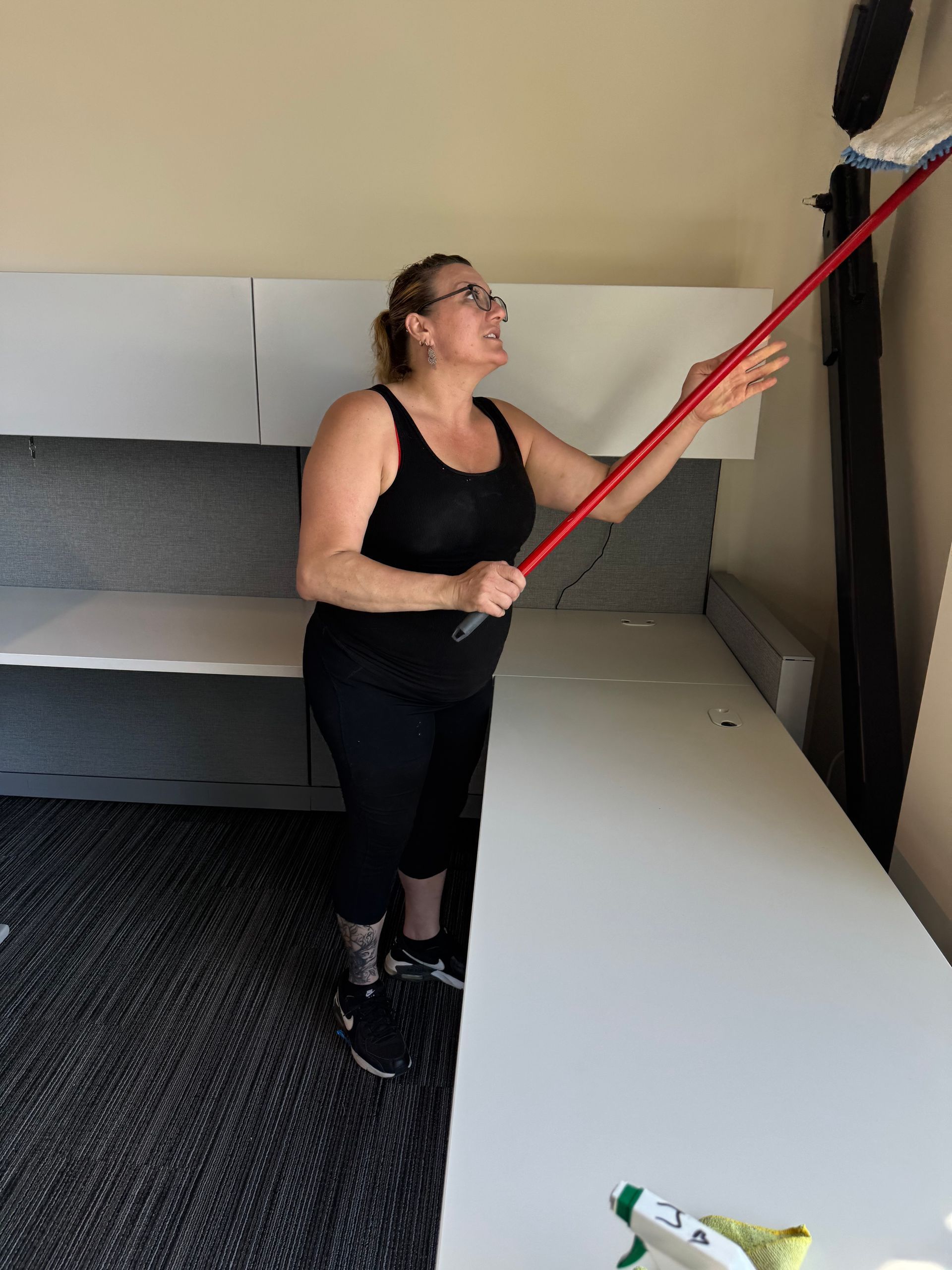 Woman in black cleaning the wall with a long-handled mop in an office setting.
