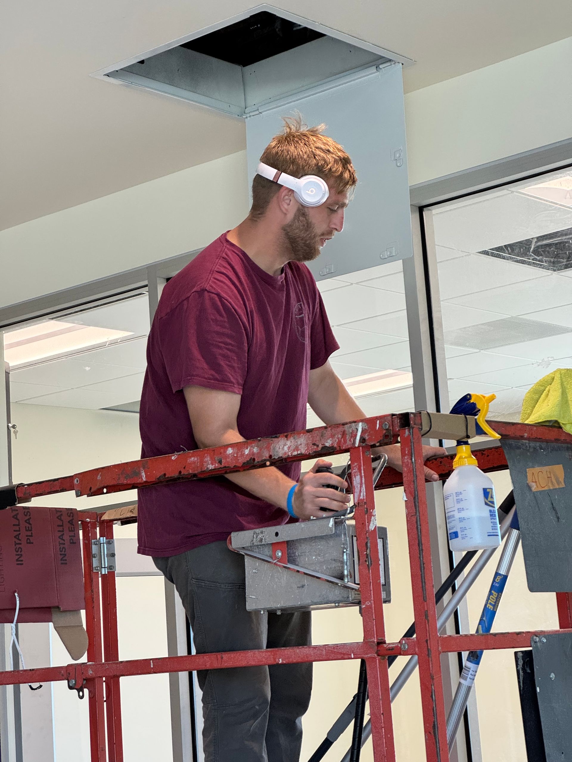 Man on a lift, working on a ceiling near a vent. He wears a maroon shirt and safety glasses.
