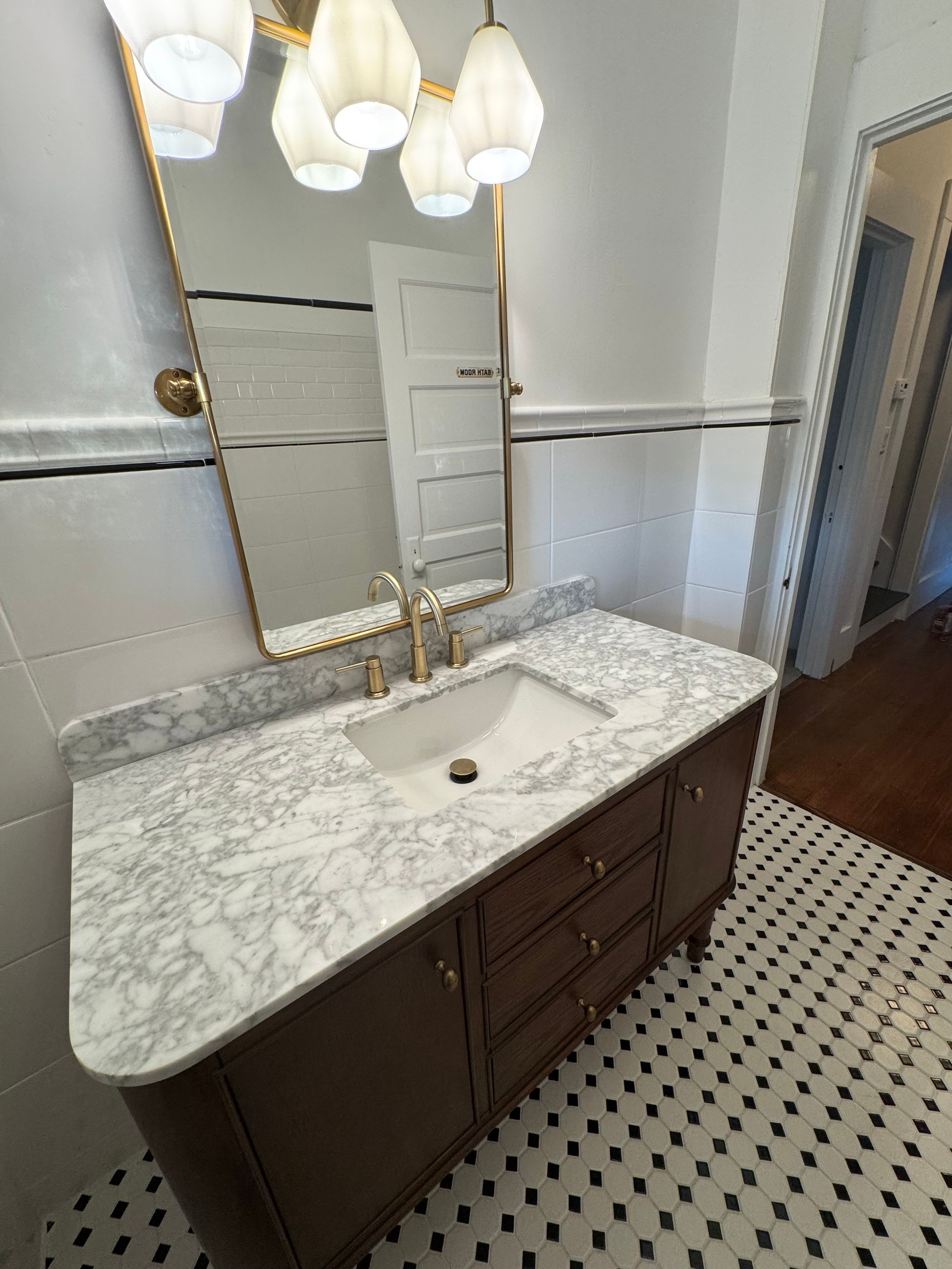 Bathroom with brown vanity, marble countertop, gold fixtures, and black and white tiled floor.
