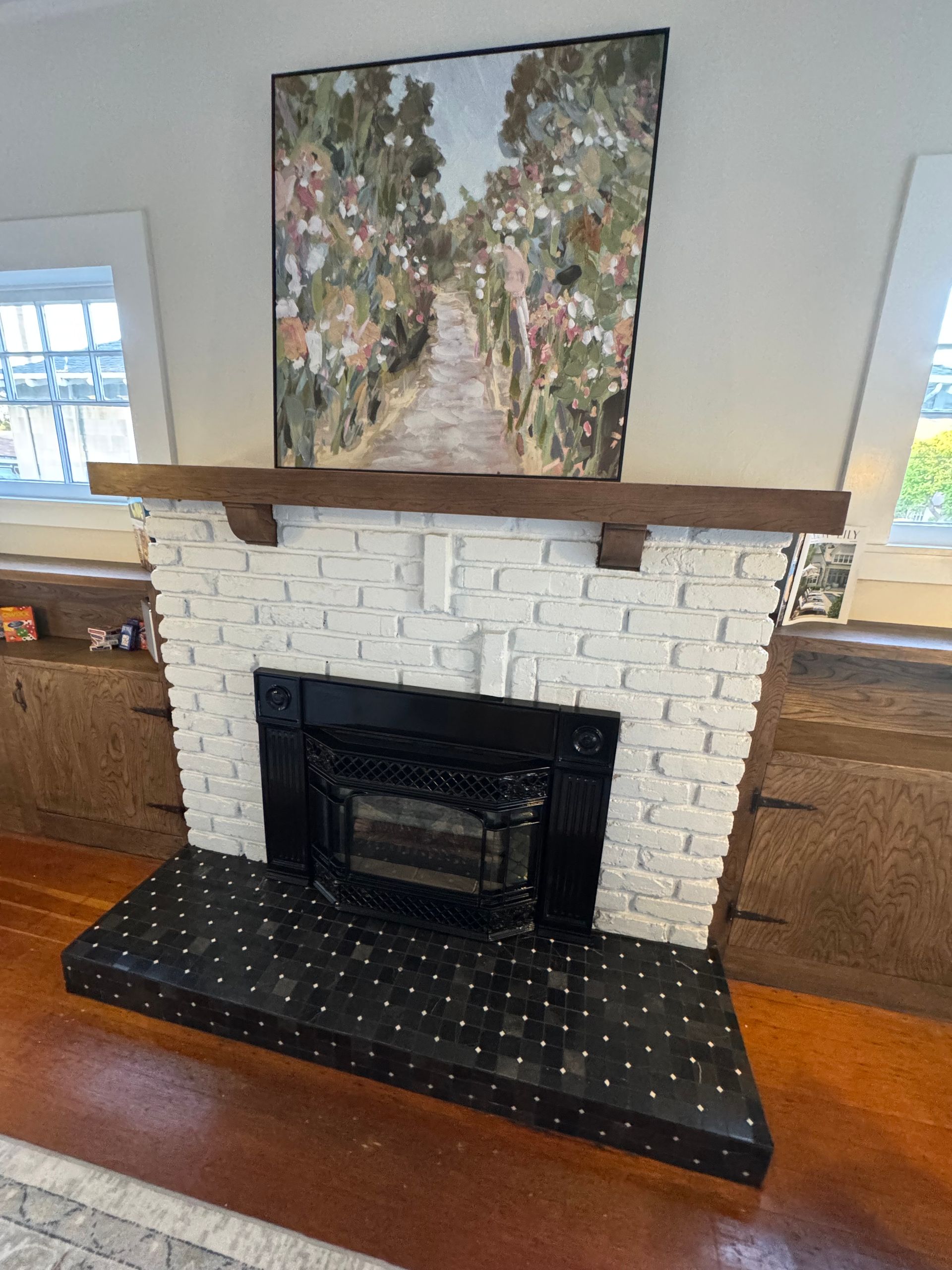 A fireplace with white brick, black hearth, and artwork of a path, set in a room with windows.