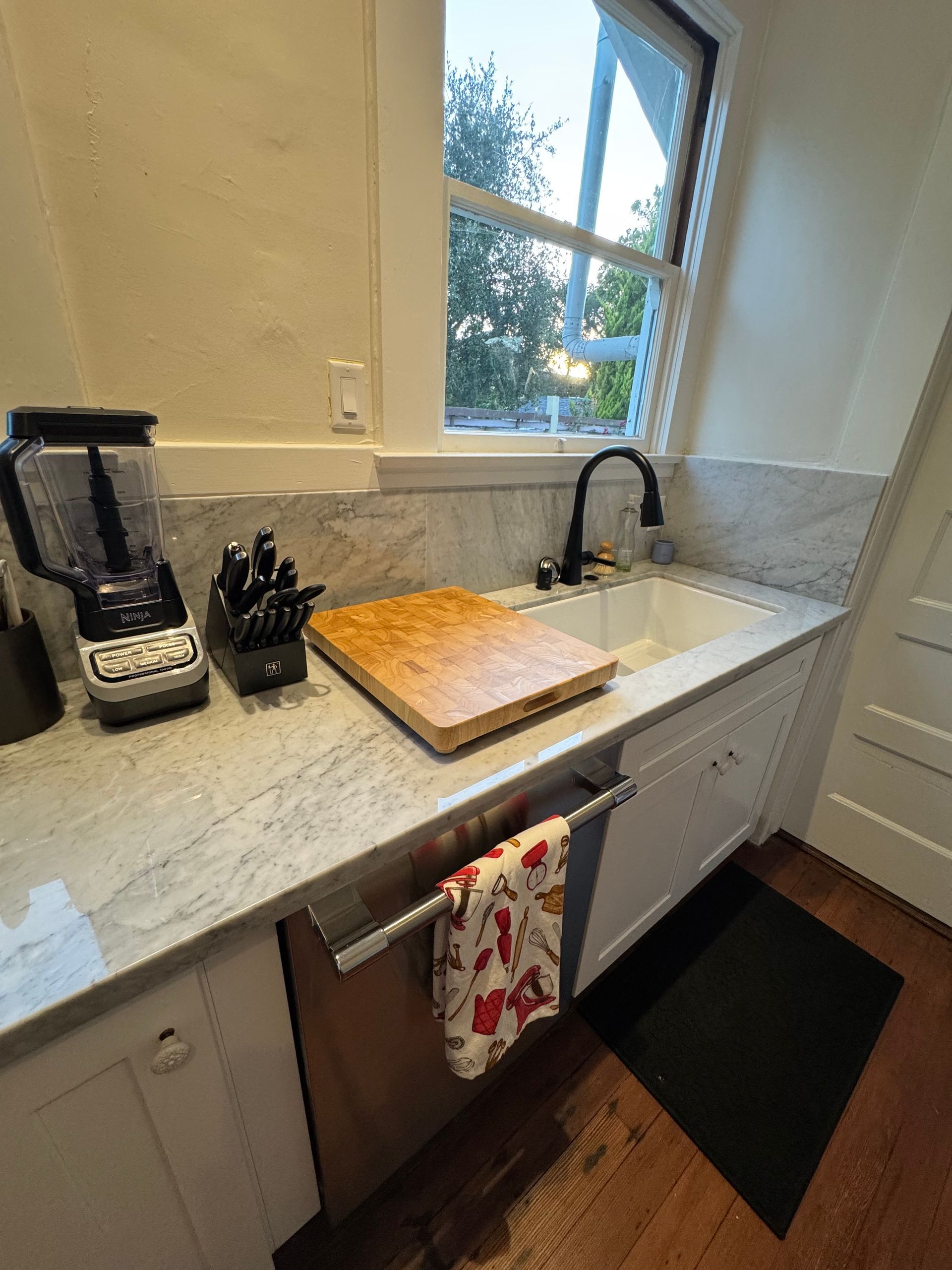 Kitchen counter with blender, knife block, cutting board, and sink near a window.