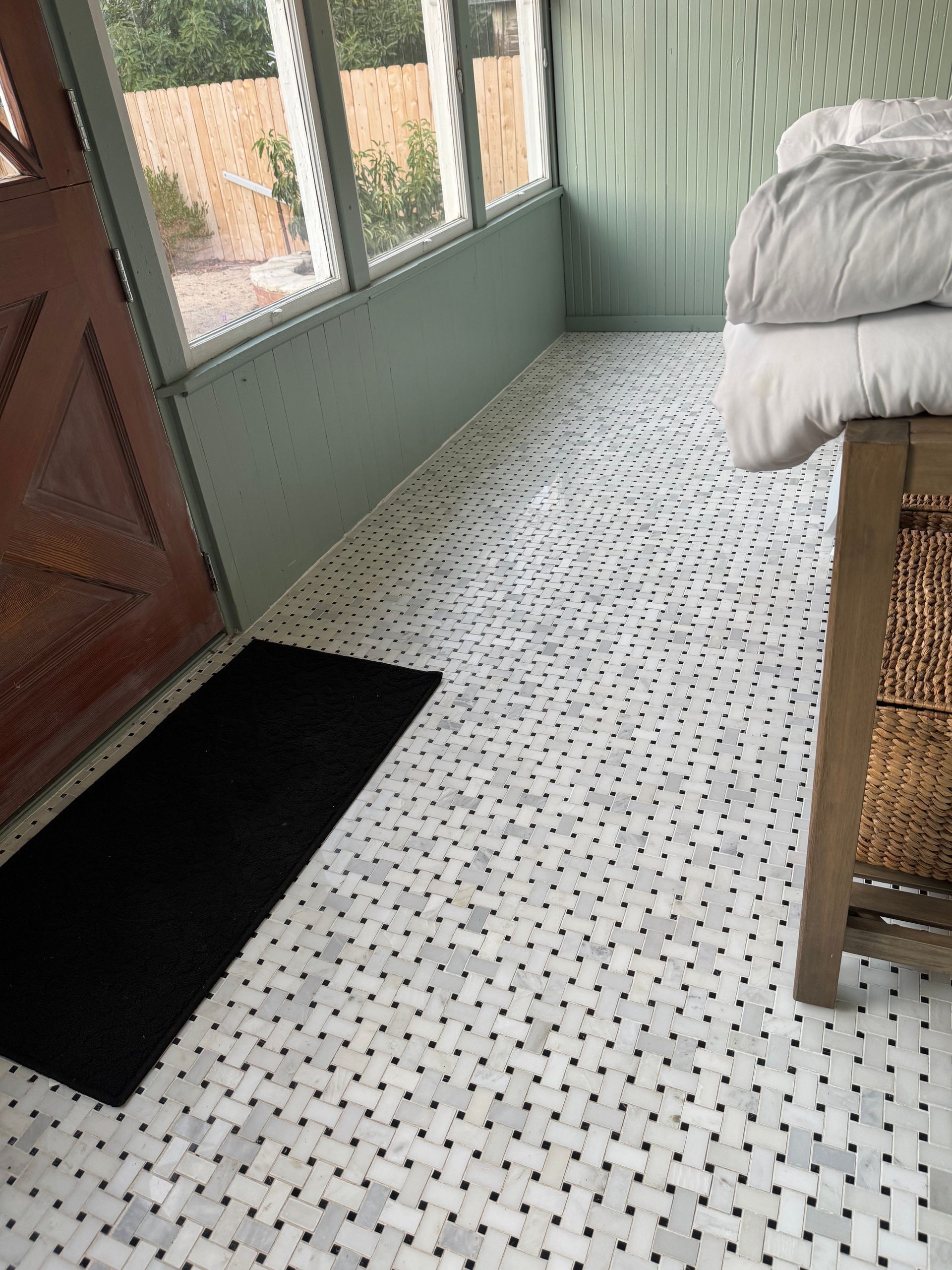 Sunroom with woven tile floor, mint walls, black rug, wooden bench, and a stack of white blankets.