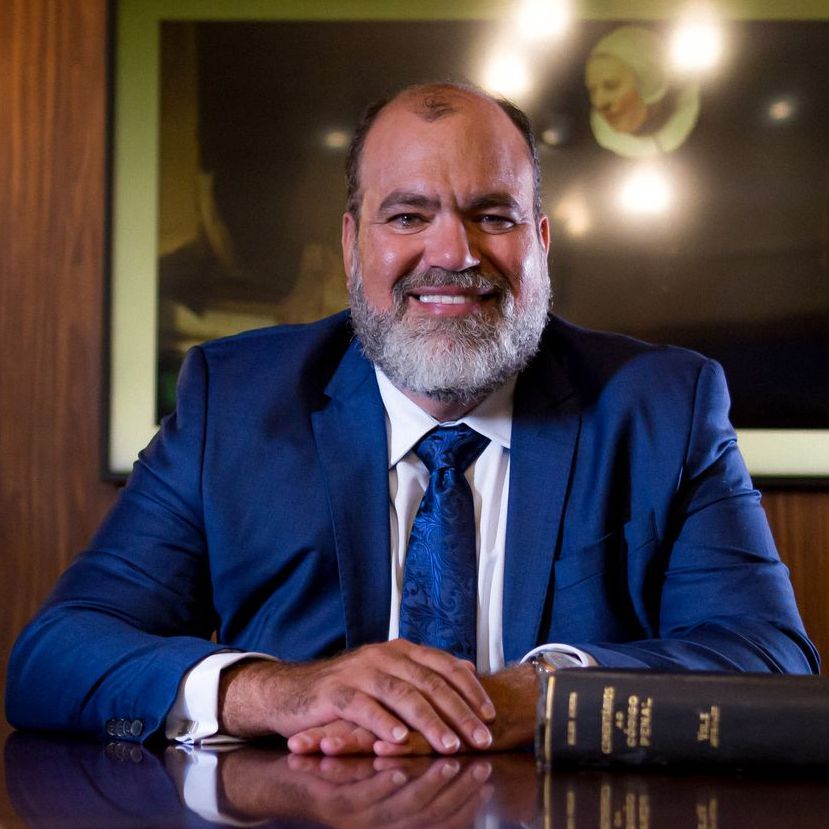 Homem de terno azul sorri, sentado a uma mesa com um livro de direito. Fundo escuro.