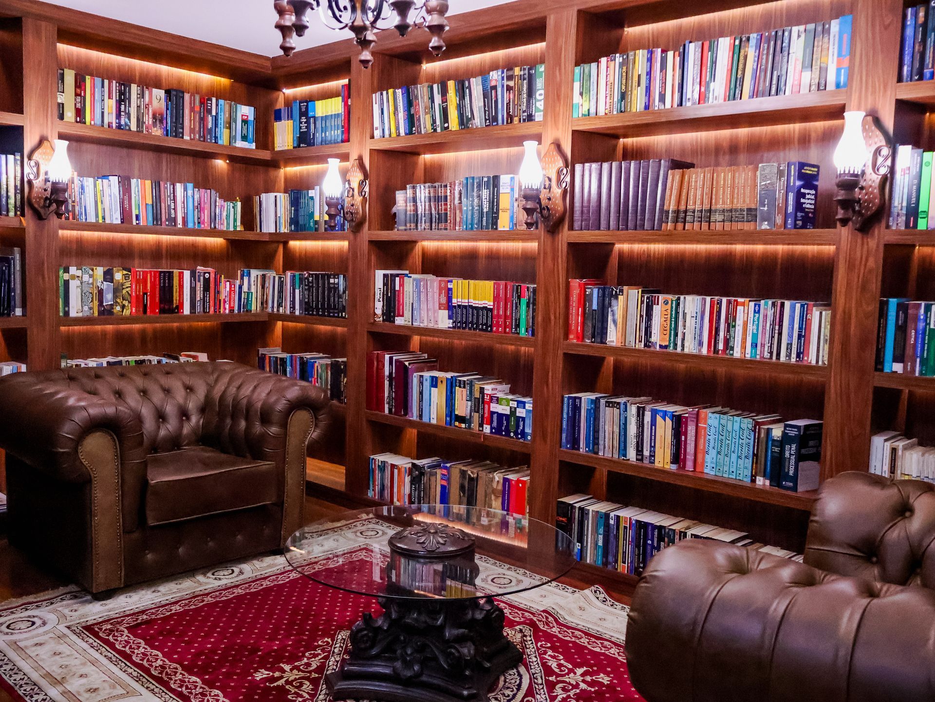 Biblioteca aconchegante com estantes de madeira escura repletas de livros, poltronas de couro e um tapete vermelho.