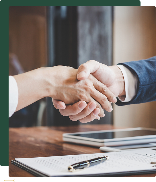 Duas pessoas apertando as mãos sobre um contrato e uma caneta em uma mesa de madeira.