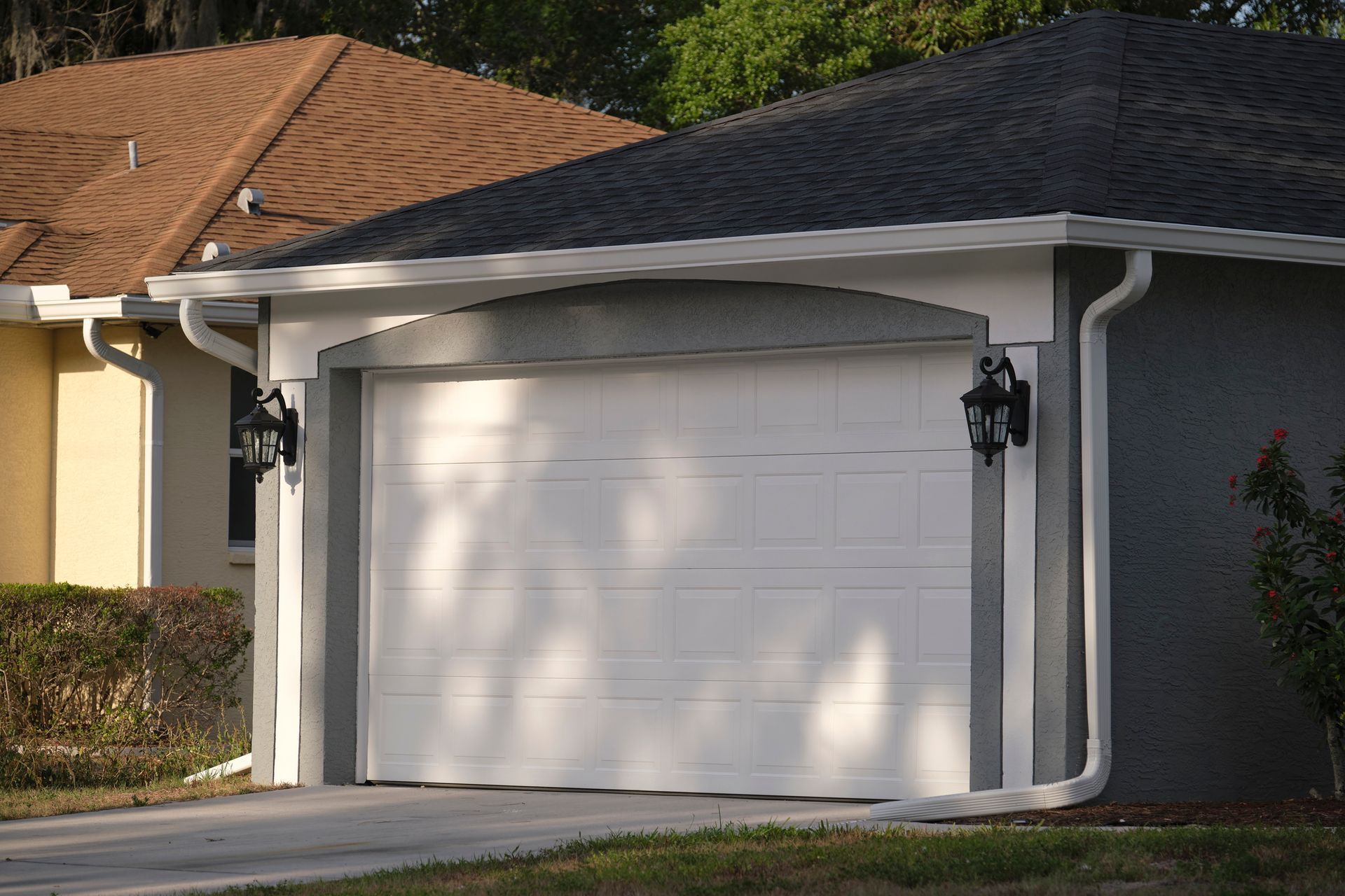 White residential garage door with exterior lamps on both sides,
rain gutters and shingle roofs.