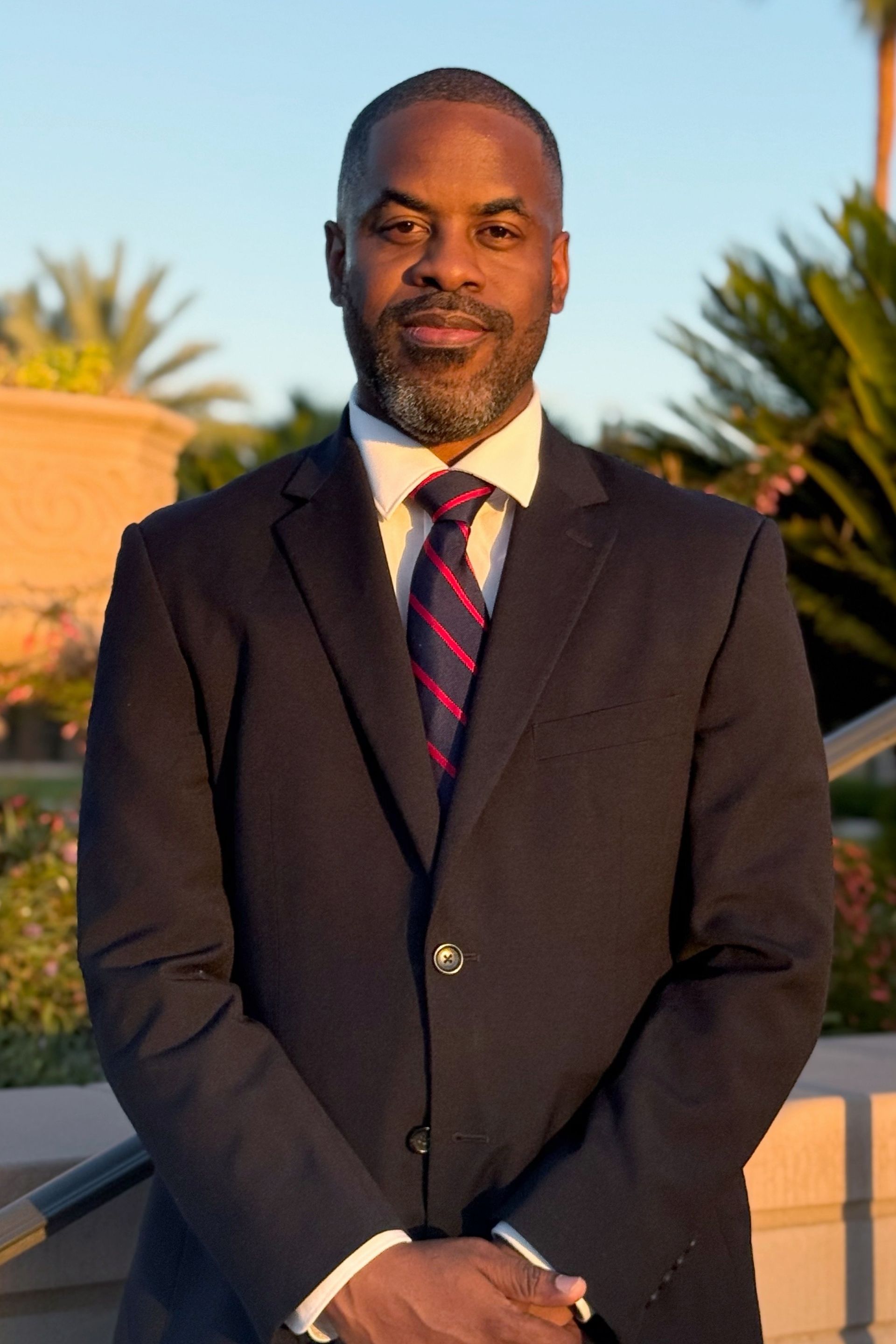 Man in a suit smiles, outdoors. Navy blazer, red striped tie, light background.