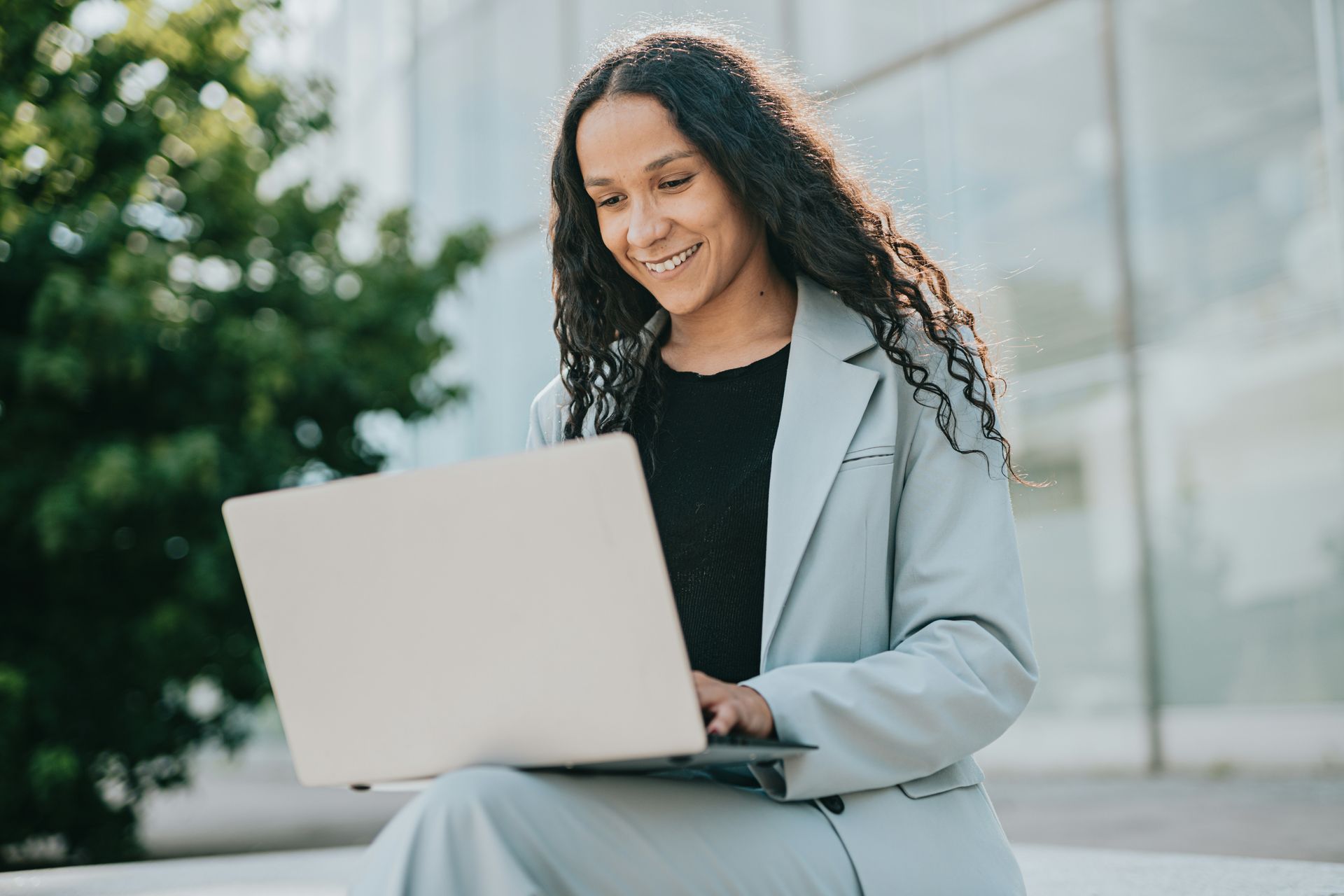 Woman in light blue suit smiles while using a laptop outside a building.