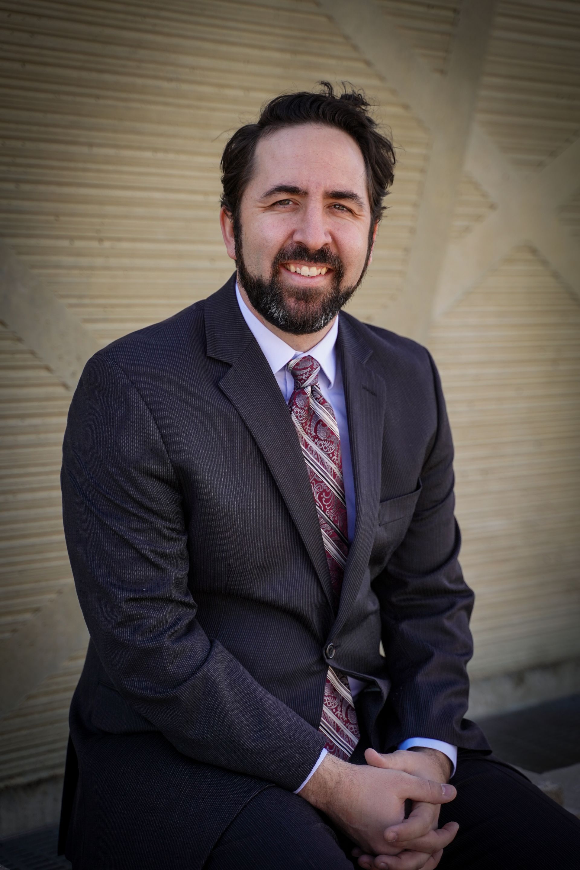 Man in suit smiling, sitting with hands clasped. He has a beard and is against a textured wall.