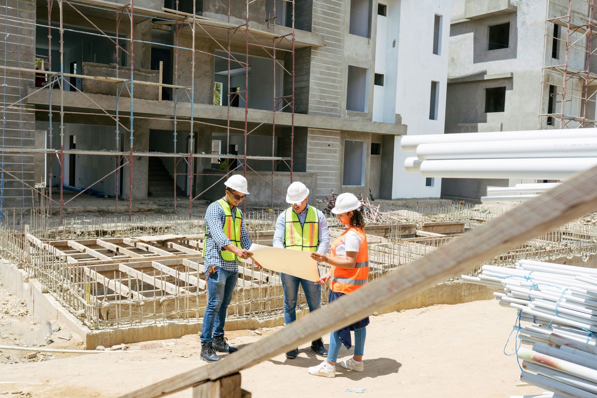 Construction workers review plans at a building site with scaffolding, cement structure, and pipes.