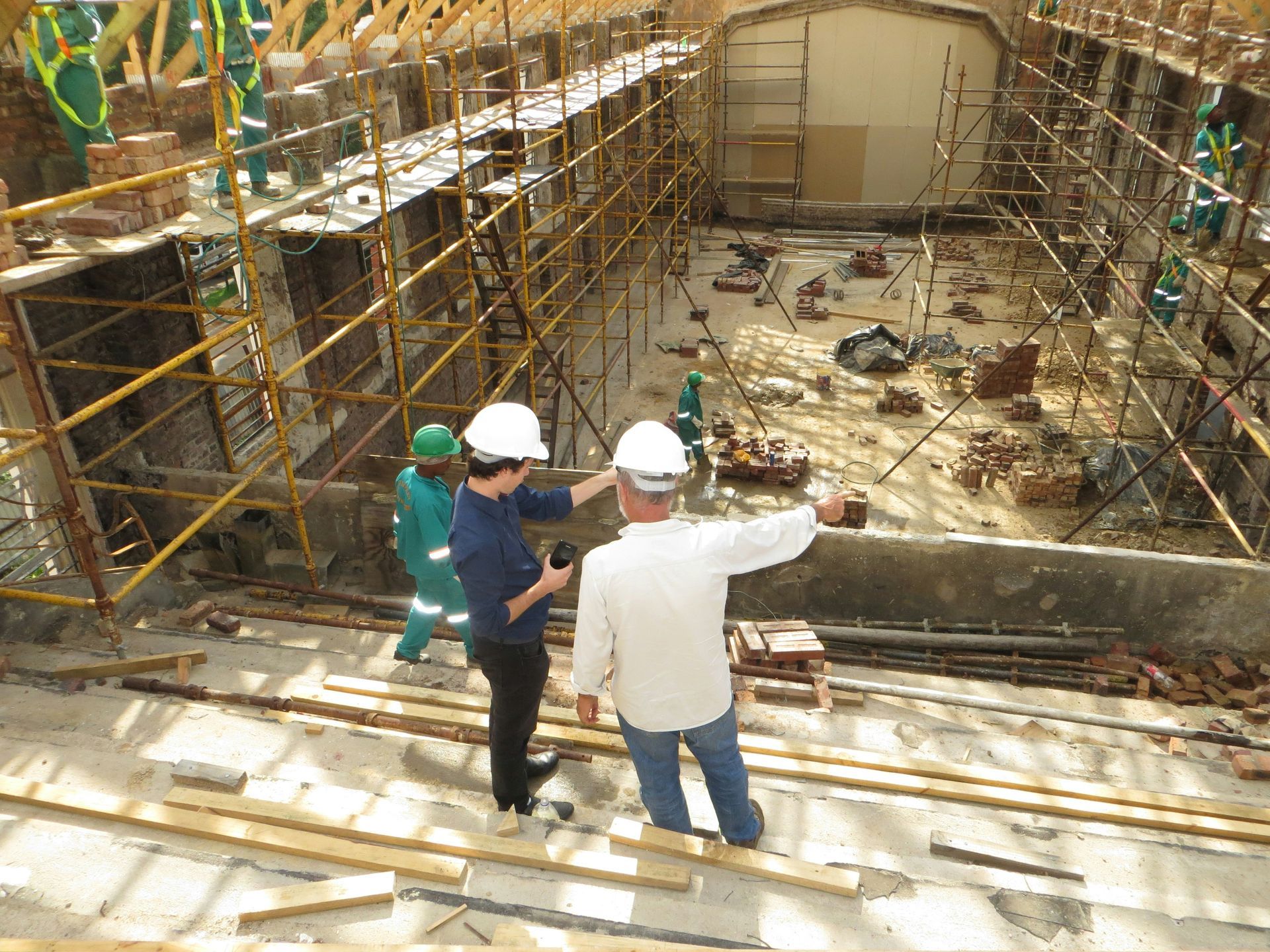 Two men in hard hats oversee construction site. Scaffolding, brickwork, and workers are visible.