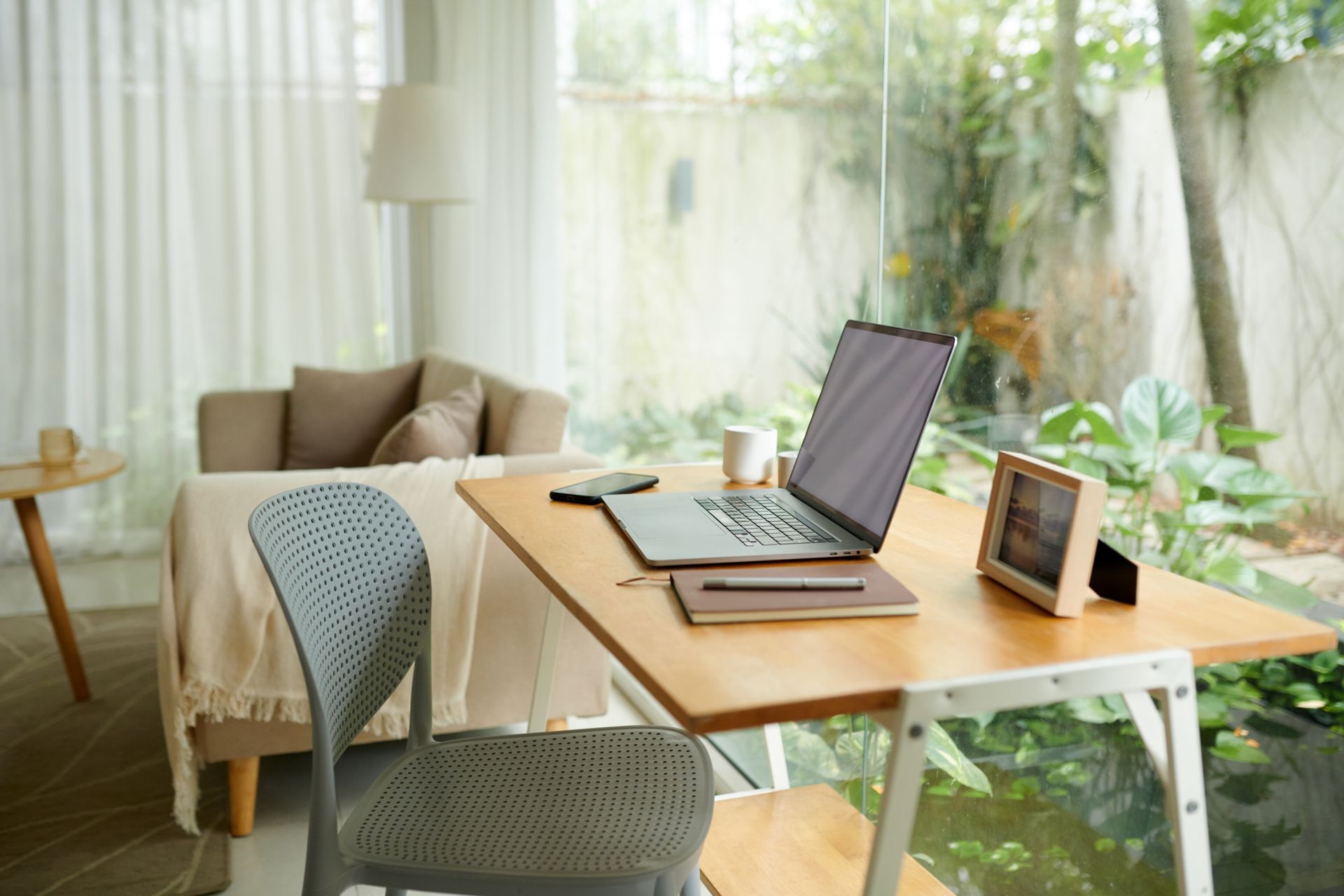 Home office with a laptop on a wooden desk. Light blue chair, sofa, and a window overlooking greenery.