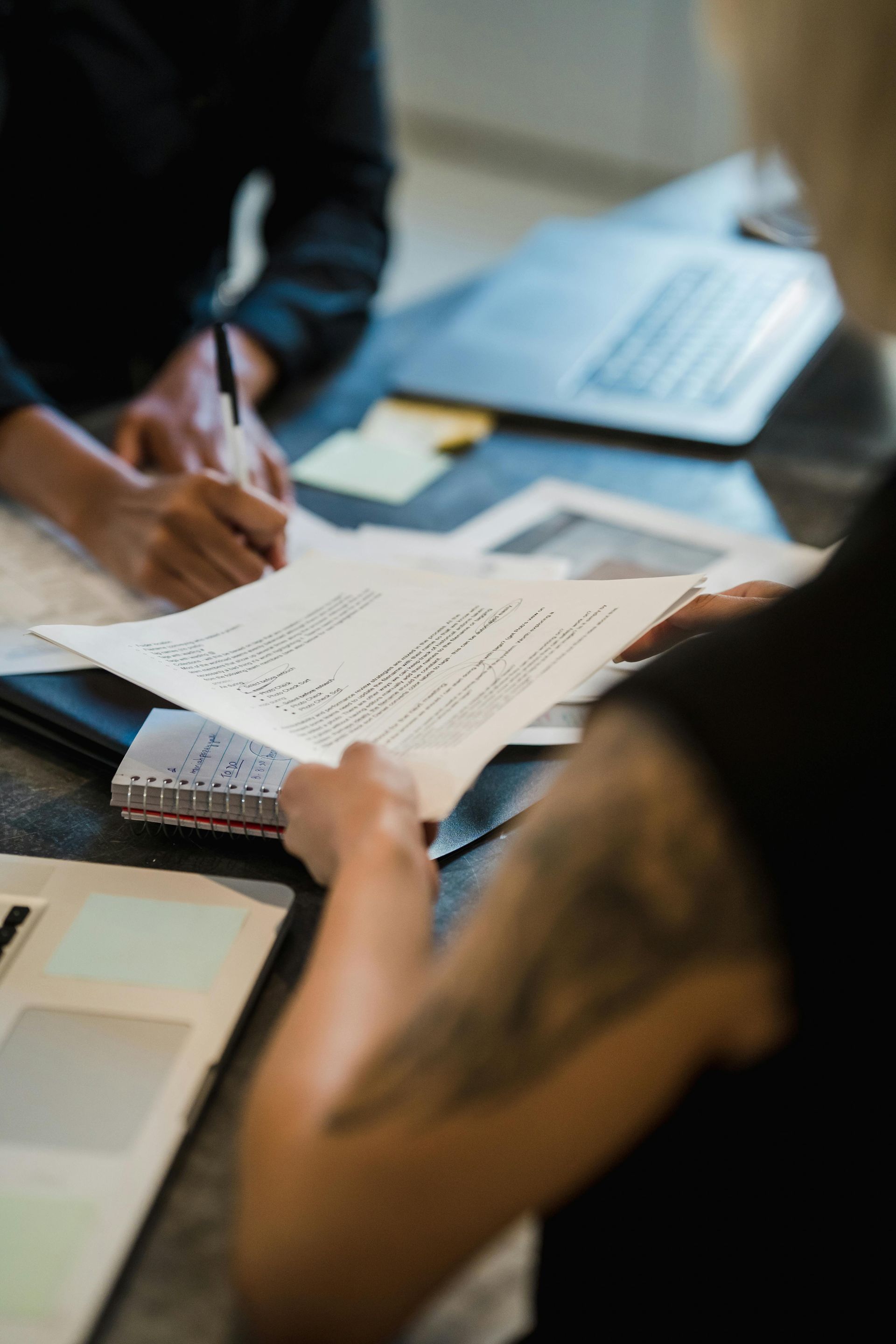 Person holding documents, reviewing paperwork with someone writing, notebook and laptop on table.