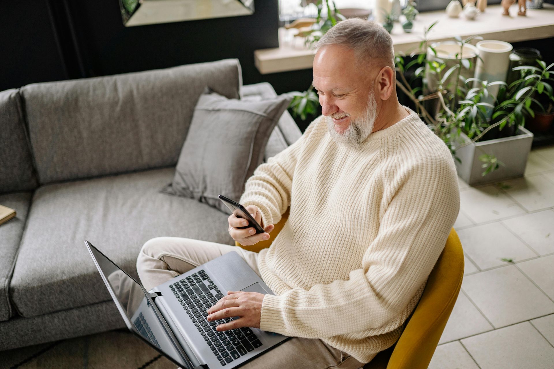 Man smiling, using a laptop and phone, seated in a chair near a sofa and plants in a room.