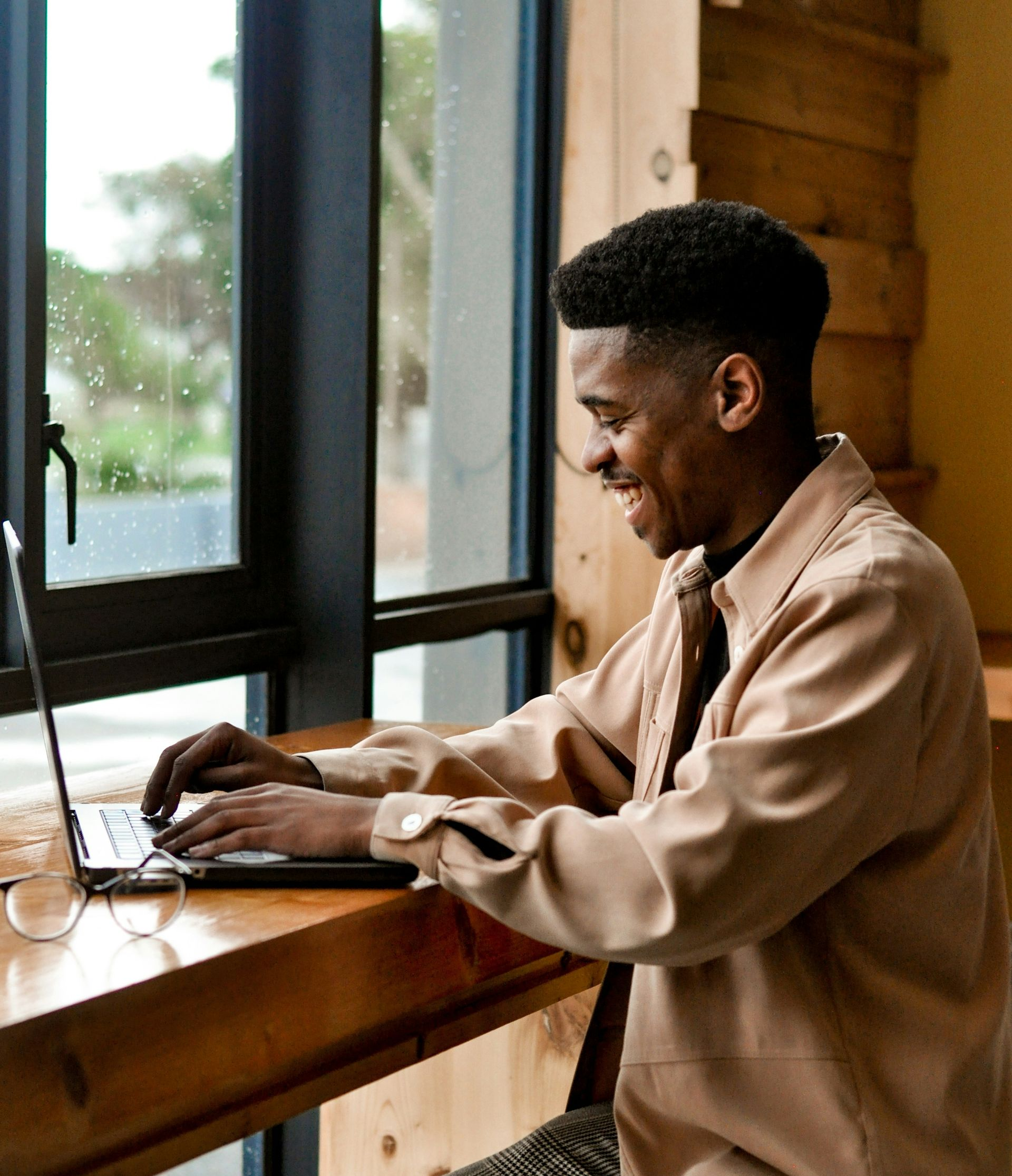 Man smiling while typing on a laptop at a wooden table near a window.