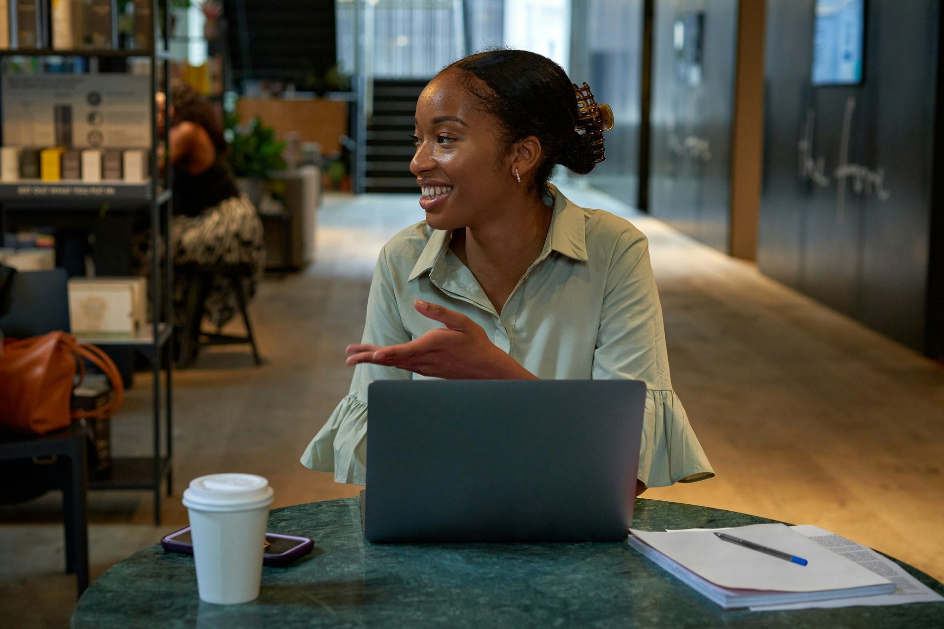 Woman smiling, gesturing, and working on a laptop at a table with coffee and notes. Inside a modern space.