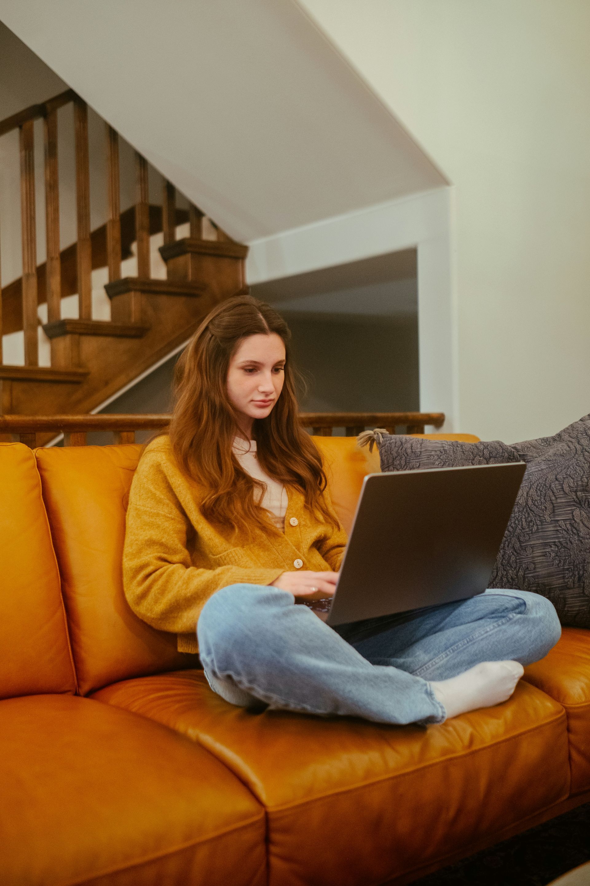 Woman in jeans and a yellow sweater using a laptop, sitting cross-legged on an orange couch.