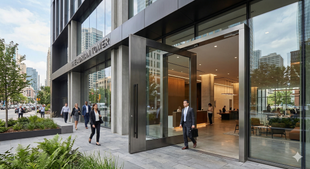 Professionals walk past the modern glass-and-steel entrance of a high-rise office building on a sunny day.