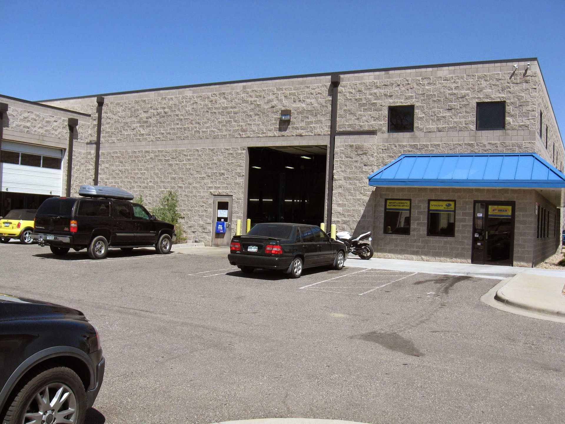 Vehicles in front of a gray brick building with a blue awning; sunny day. | 5th Gear Automotive