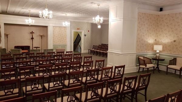 Rows of brown chairs face a room with chandeliers and a cross, likely a chapel or memorial hall.