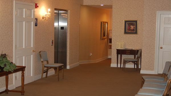 Hallway with elevator and chairs, tan walls, brown carpet, and two framed pictures.