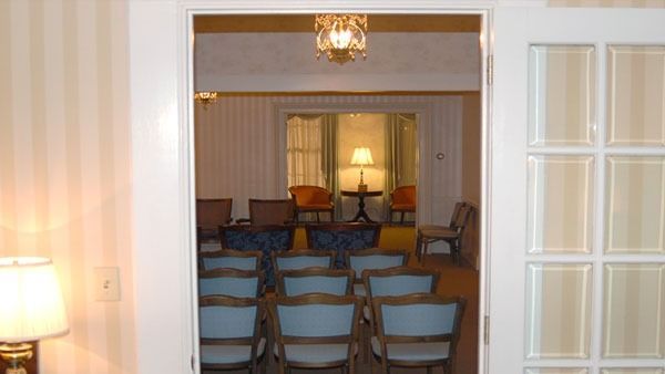 Interior with chairs arranged in rows facing a table and two orange chairs, seen through a doorway.
